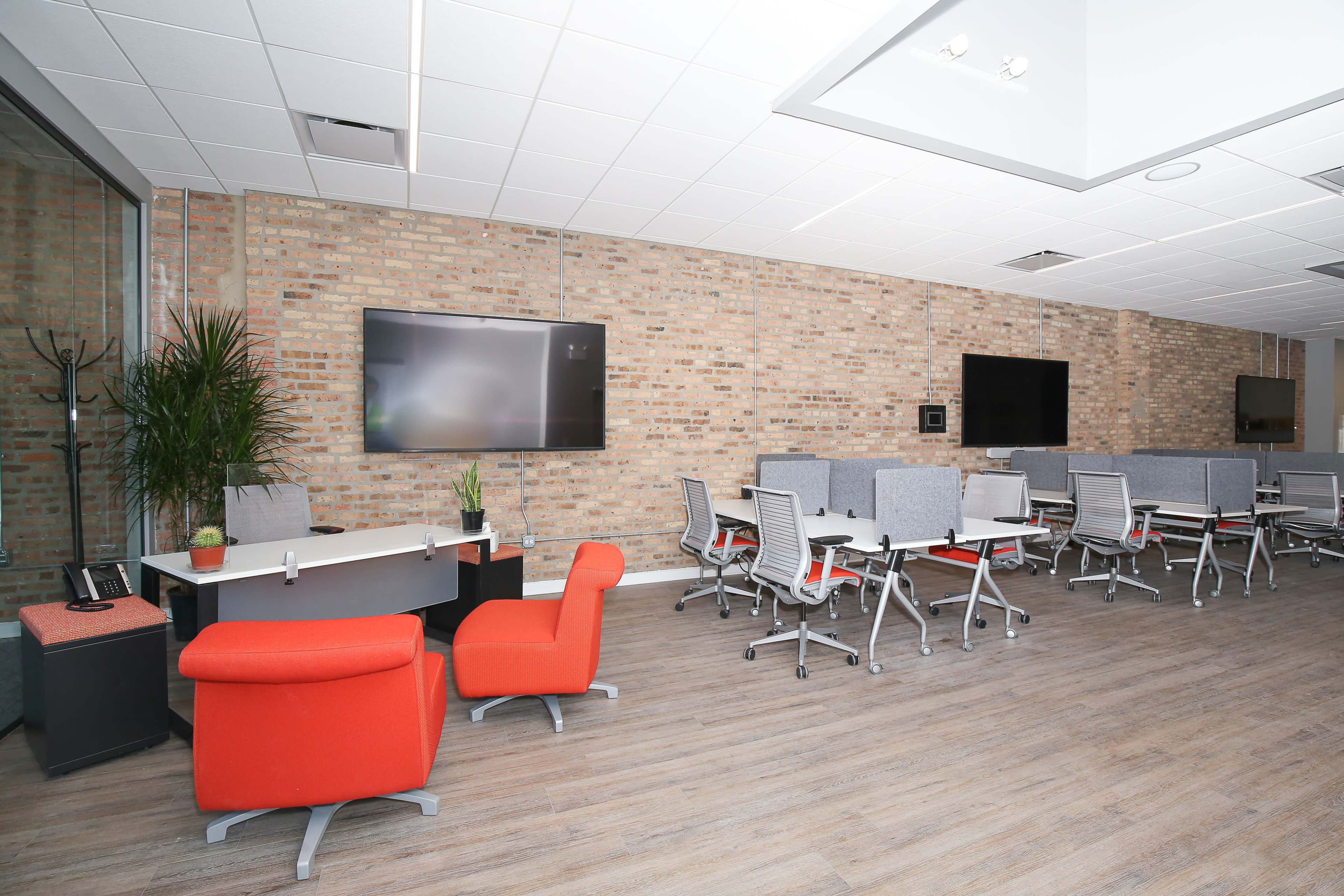 A modern office space with a brick wall, a reception desk, orange chairs, and several workstations equipped with screens and ergonomic chairs.