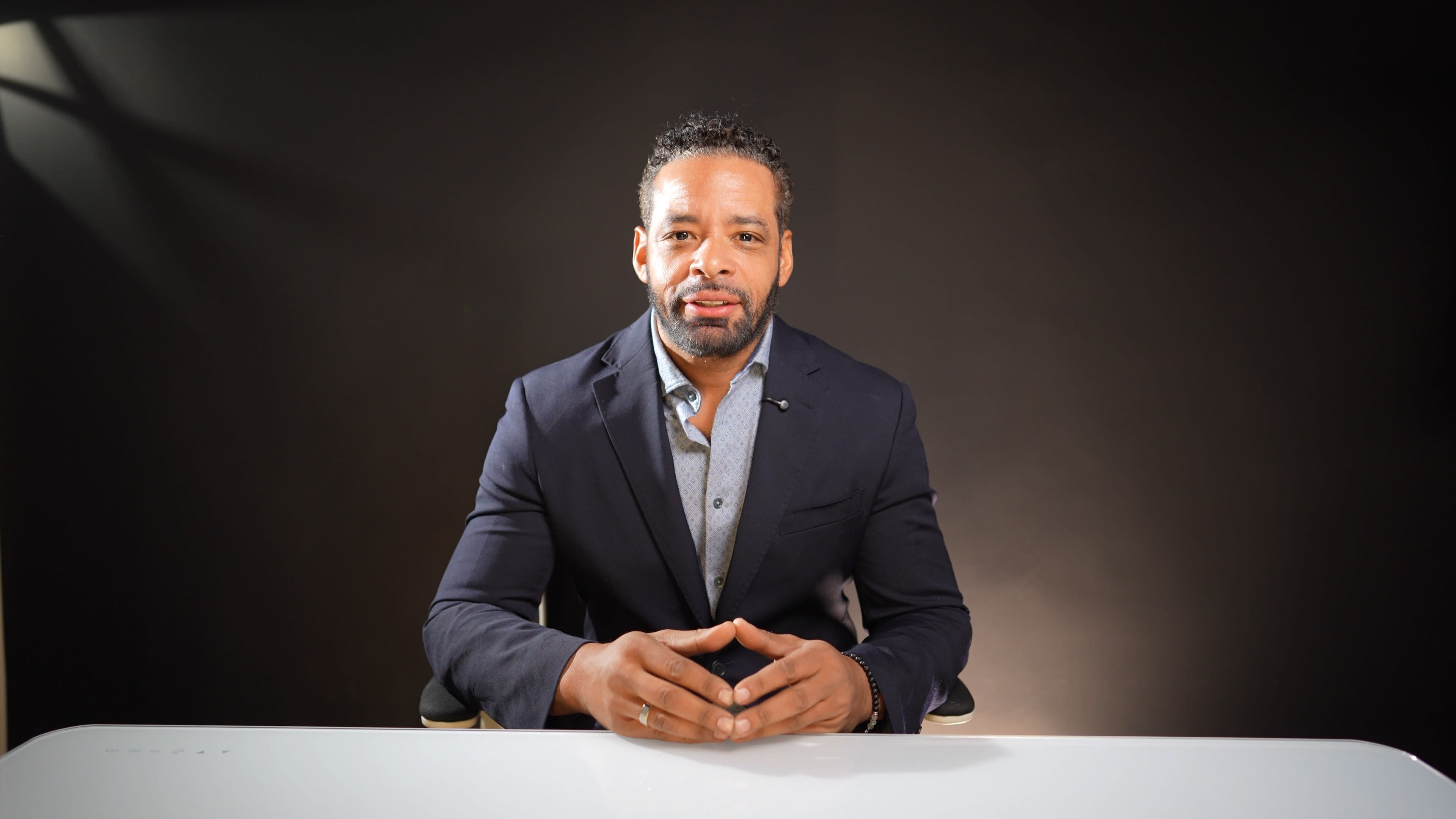 A man in a dark suit sits at a white table with his hands clasped, facing the camera against a dark background.