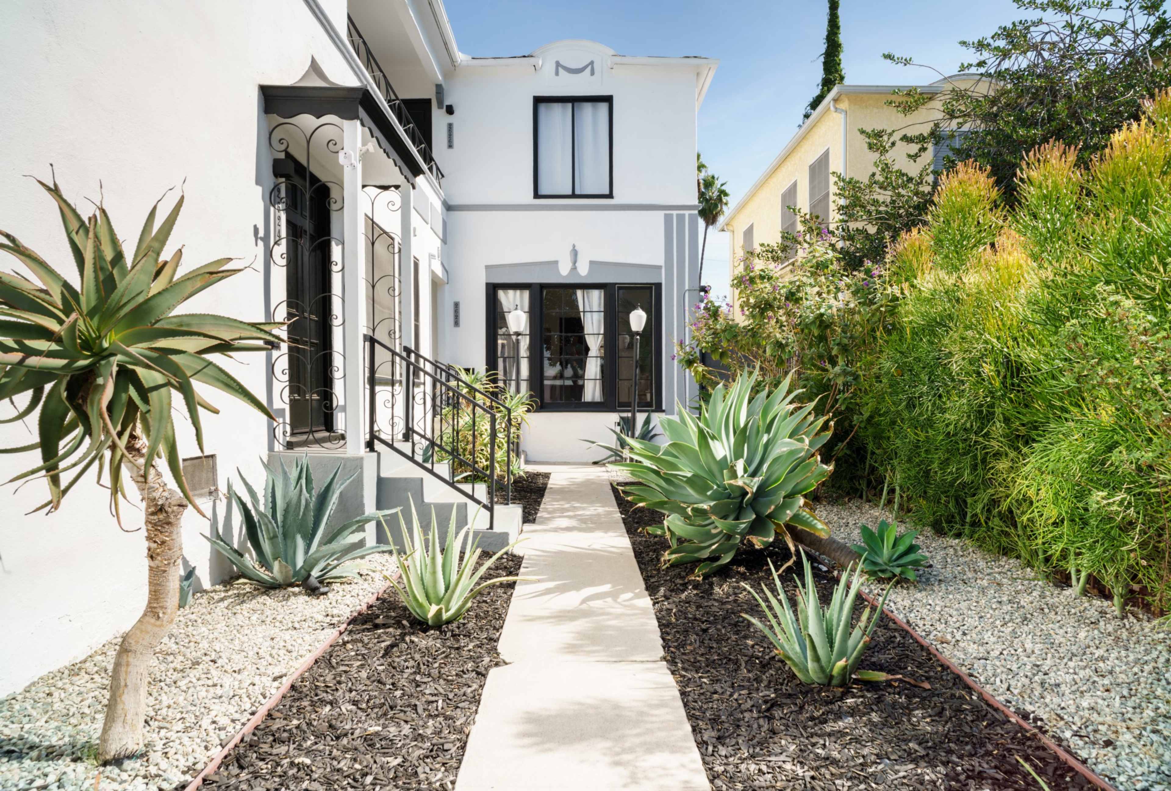 The image shows a pathway leading to a white house, bordered by decorative plants and gravel landscaping on either side.