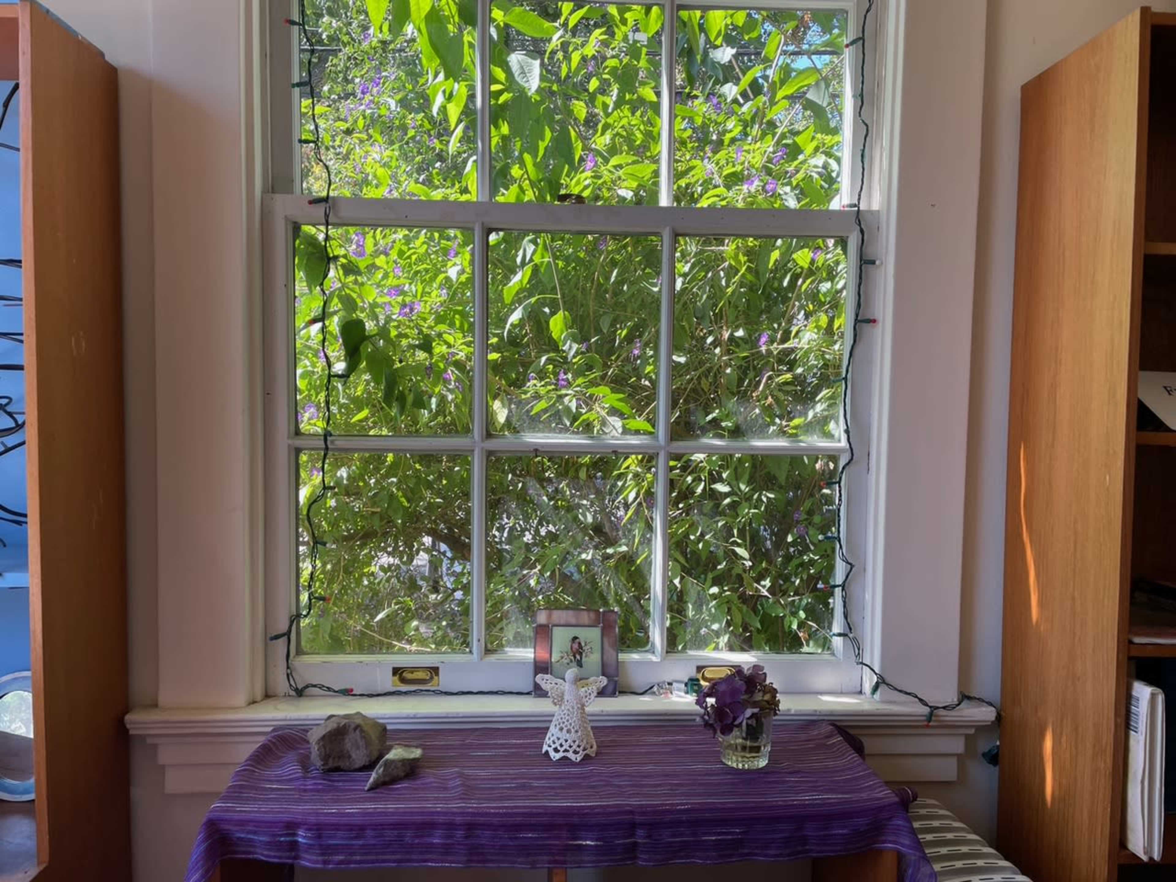 A sunlit window surrounded by greenery overlooks a small table with a decorative figure and a vase of flowers.