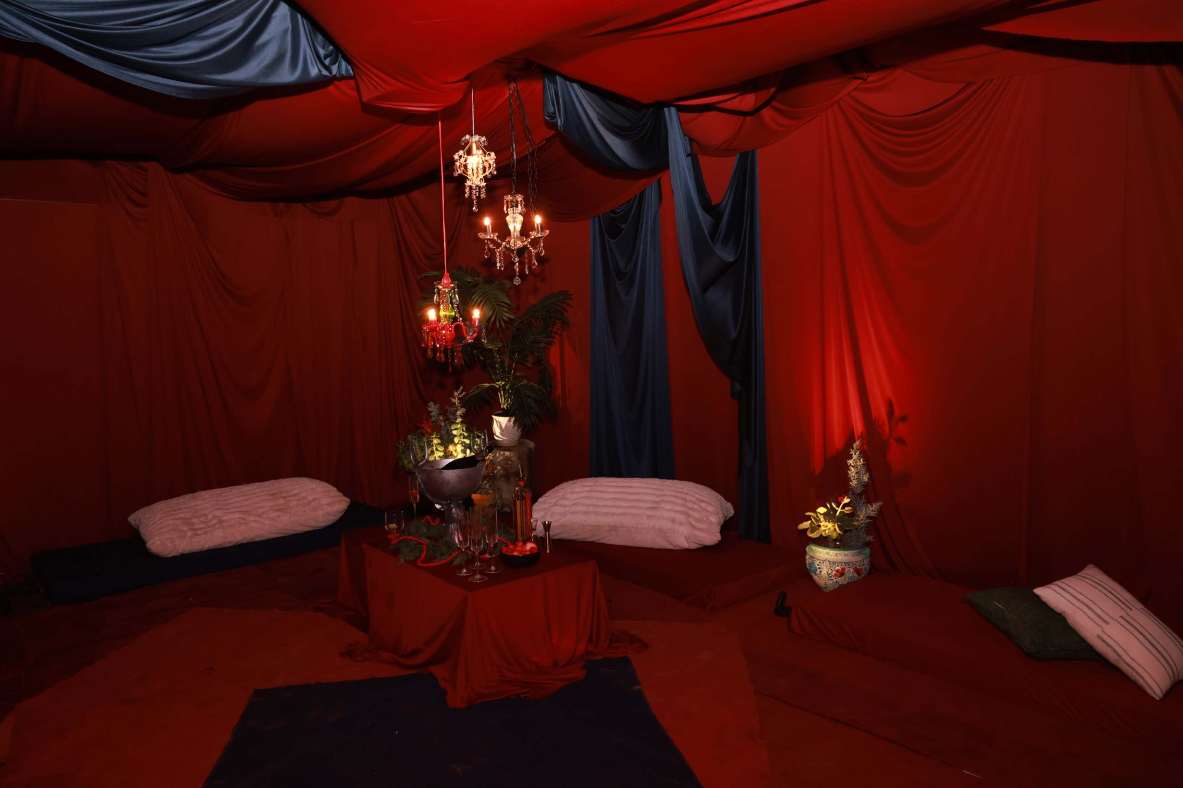 The image shows a dimly lit, red-themed tent interior with draped fabric, a low table adorned with decorative items, and cushioned seating on the floor.