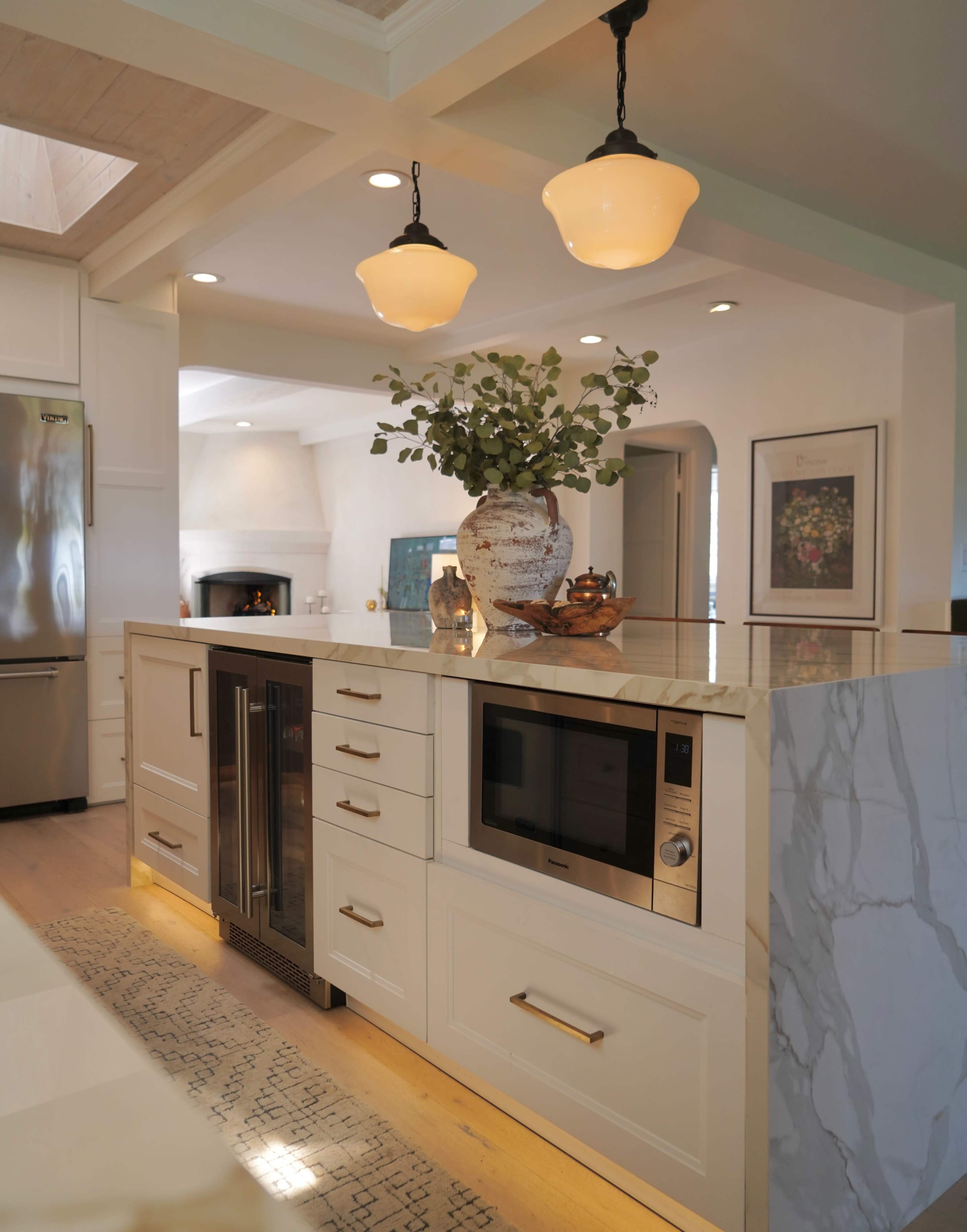 The image shows a modern kitchen with a central island featuring a microwave, wine cooler, and decorative vase, illuminated by pendant lights.
