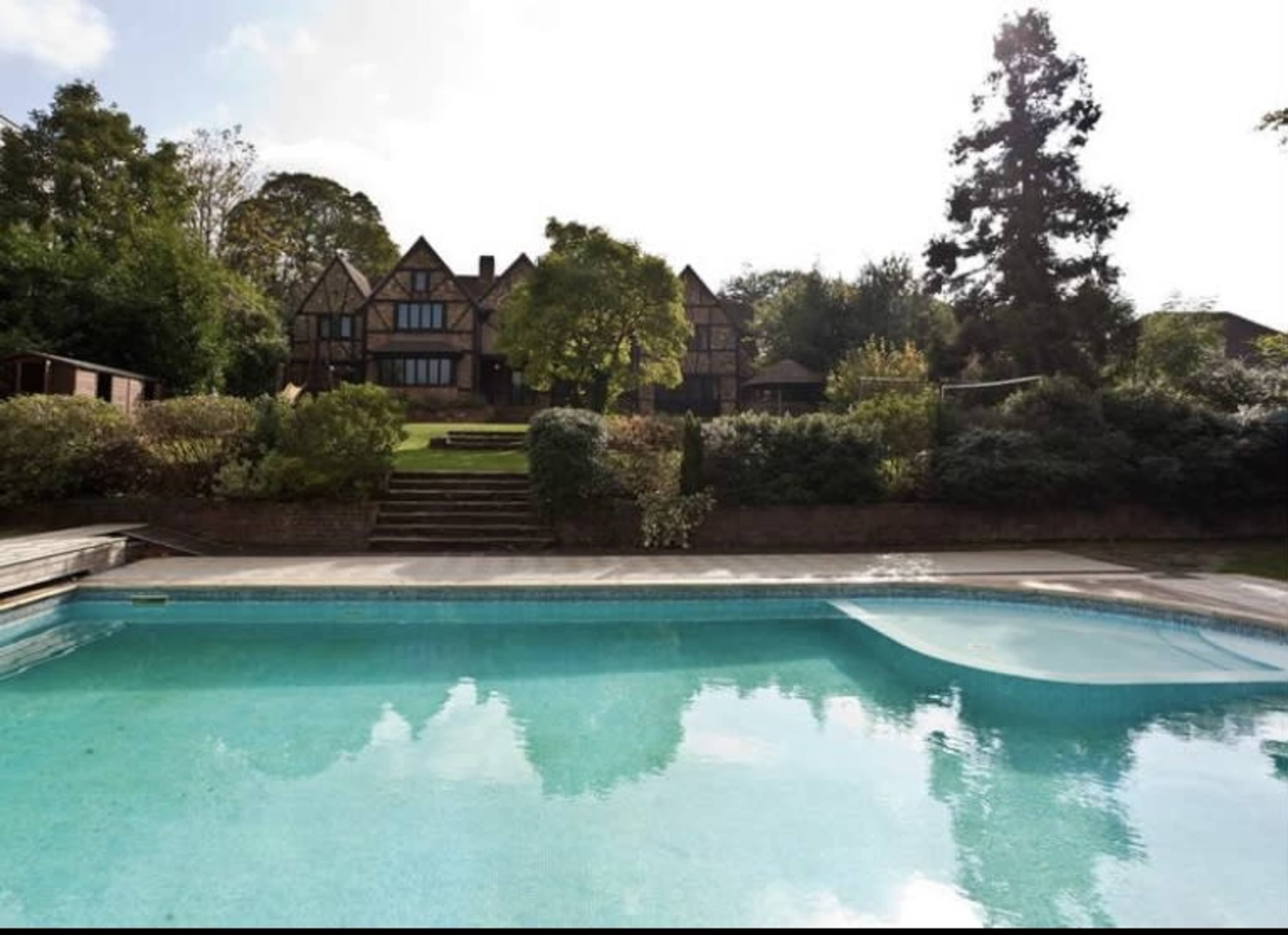 A large, traditional-style house is visible in the background, set on a hillside next to a swimming pool surrounded by landscaped greenery.
