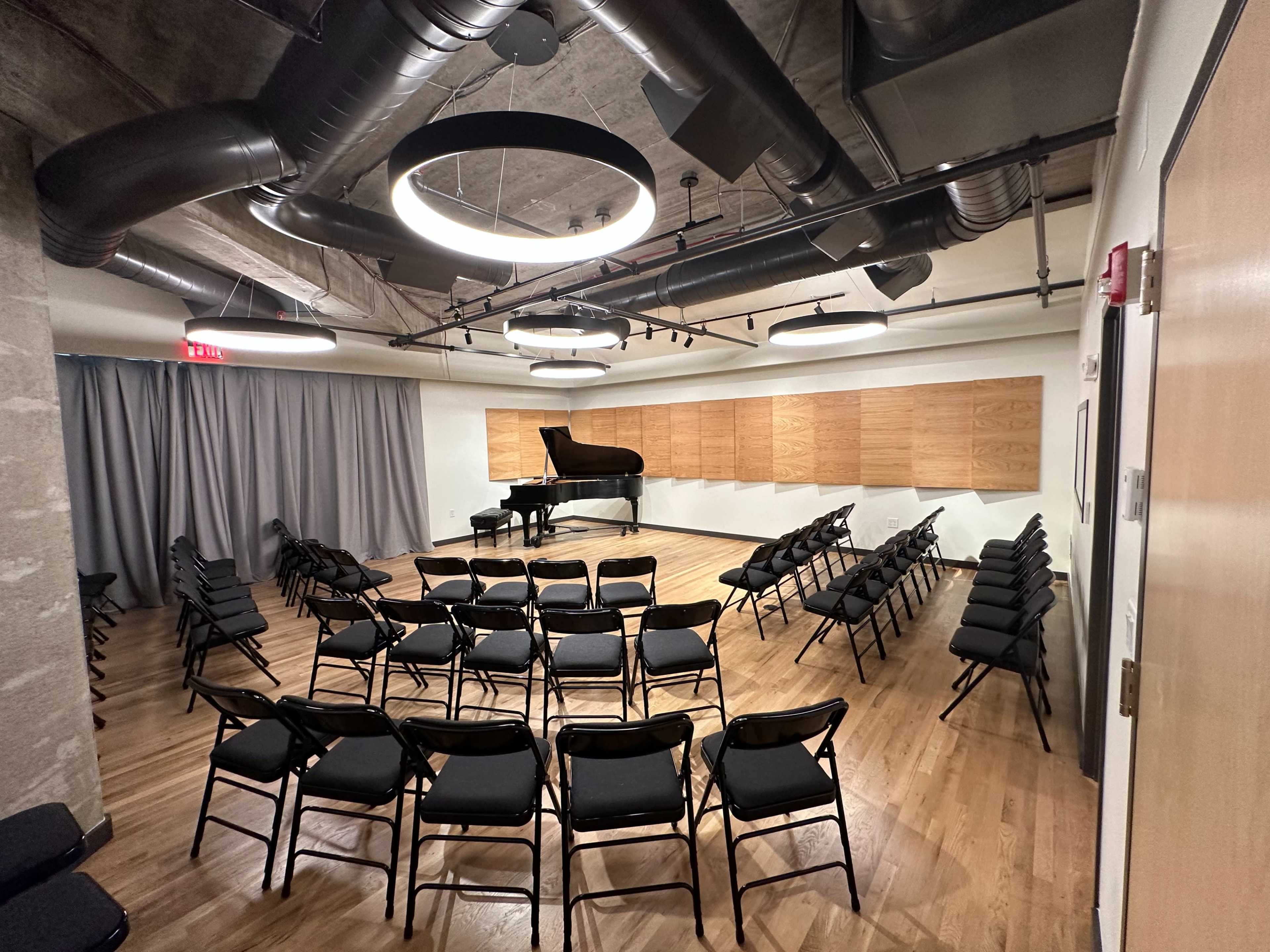A performance space with a grand piano on a wooden stage, surrounded by rows of black folding chairs and acoustic wall panels.