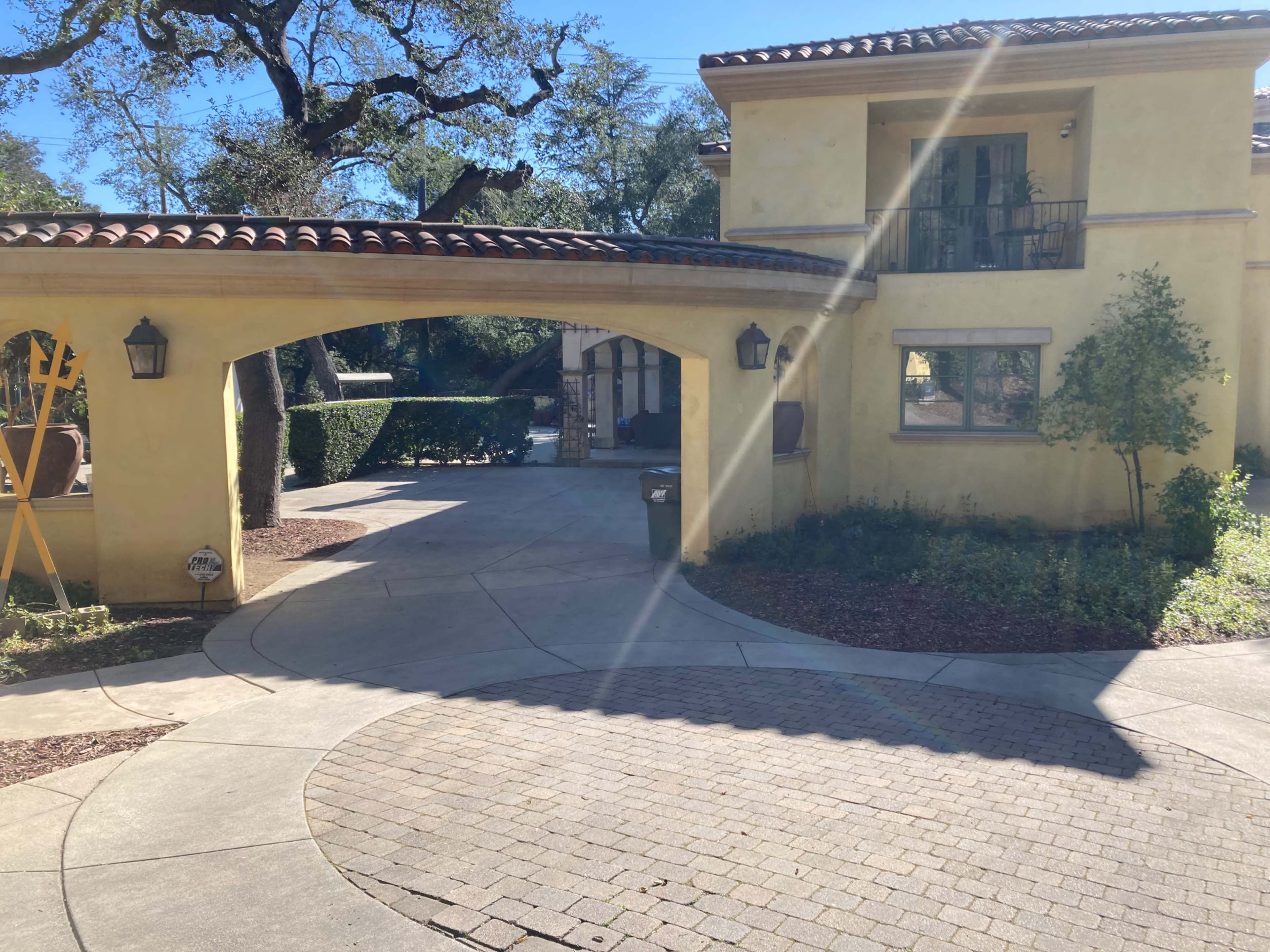 A yellow stucco house with a tiled roof is situated beside a circular driveway bordered by greenery and stone pathways.