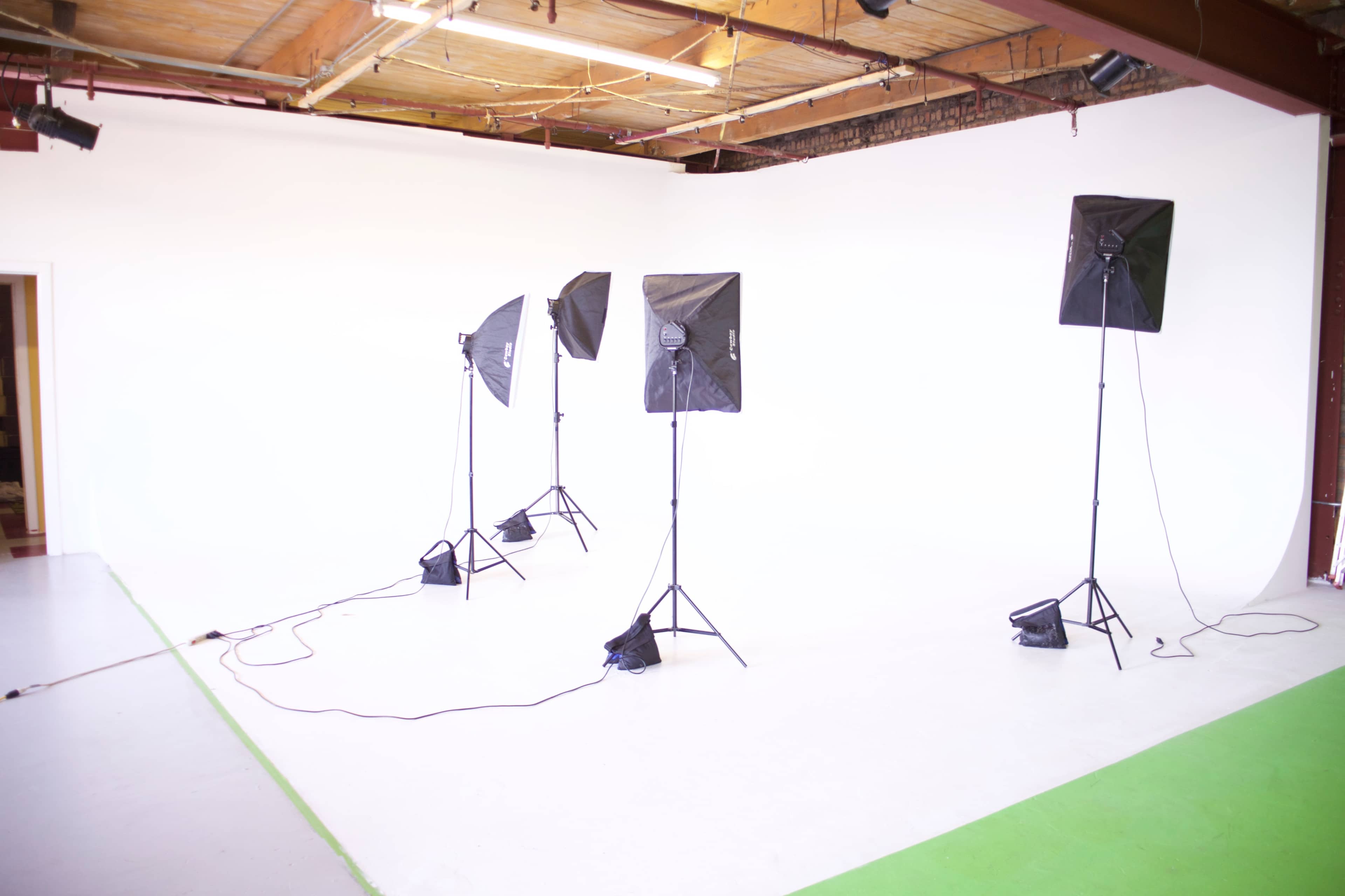 A photo studio with four black softbox lights set up on tripods in front of a white backdrop and a section of green flooring.