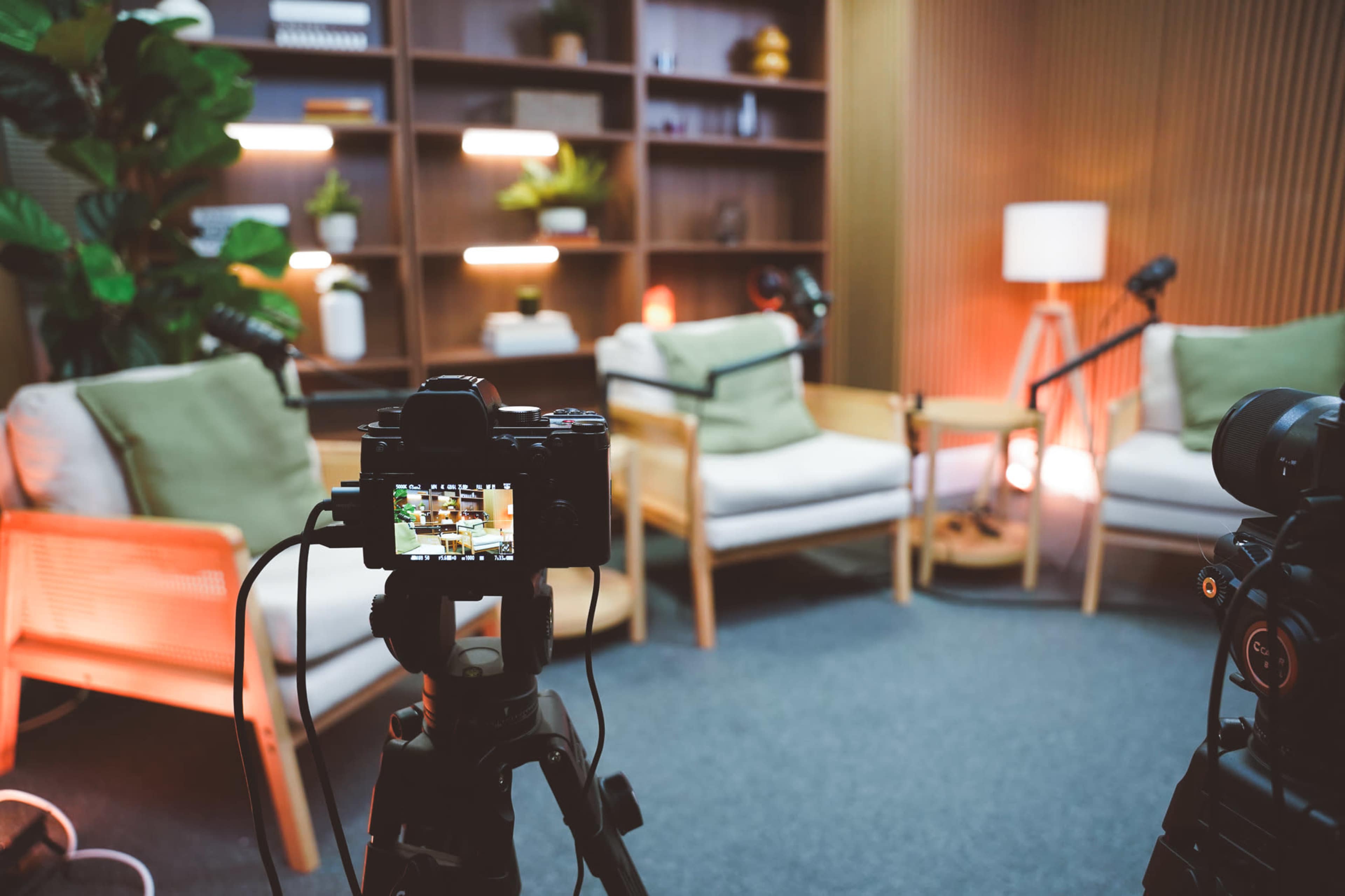 A camera is focused on a cozy studio setup with two armchairs, a small table, and plants arranged on shelves in the background.
