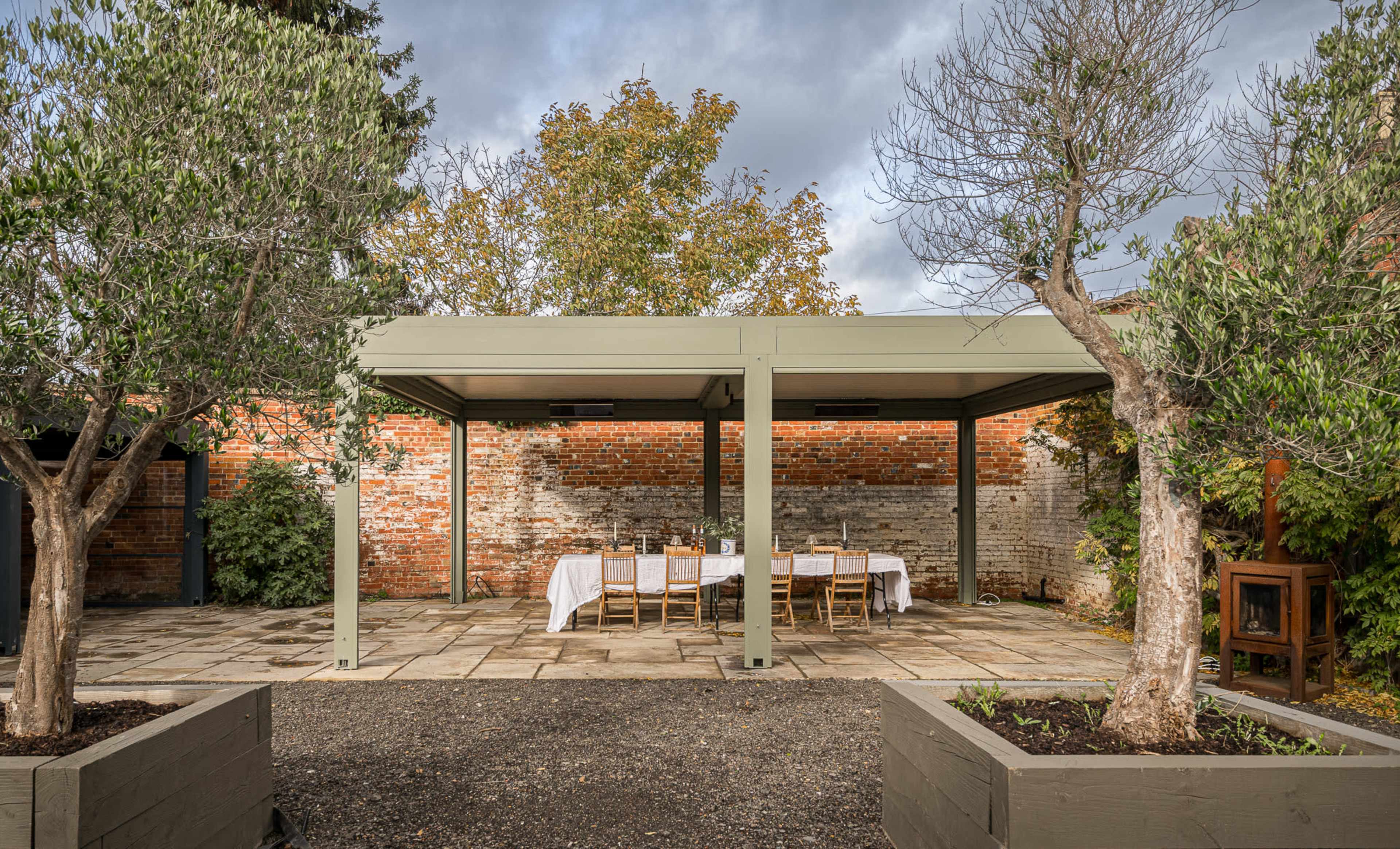 An outdoor dining area with a long table and chairs is set under a wooden gazebo, surrounded by trees and brick walls.