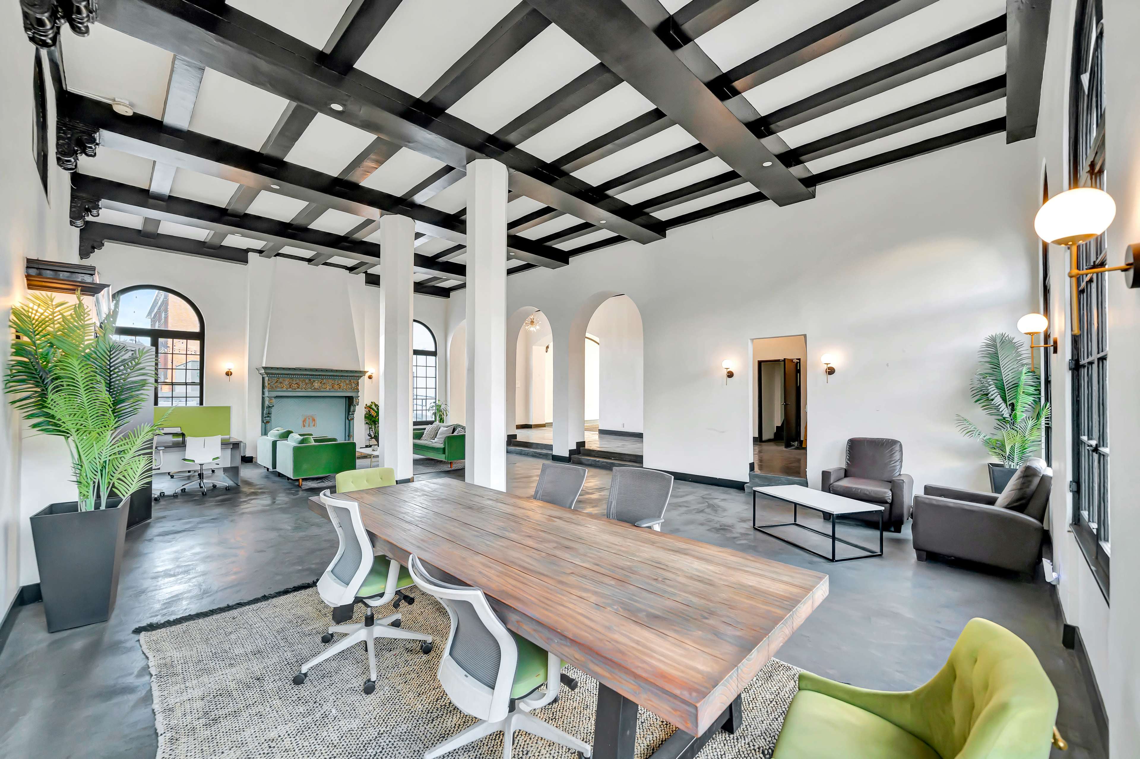 The image shows a spacious, modern office interior with exposed wooden beams, a large wooden table surrounded by chairs, and light-colored walls, featuring various seating areas and decorative plants.