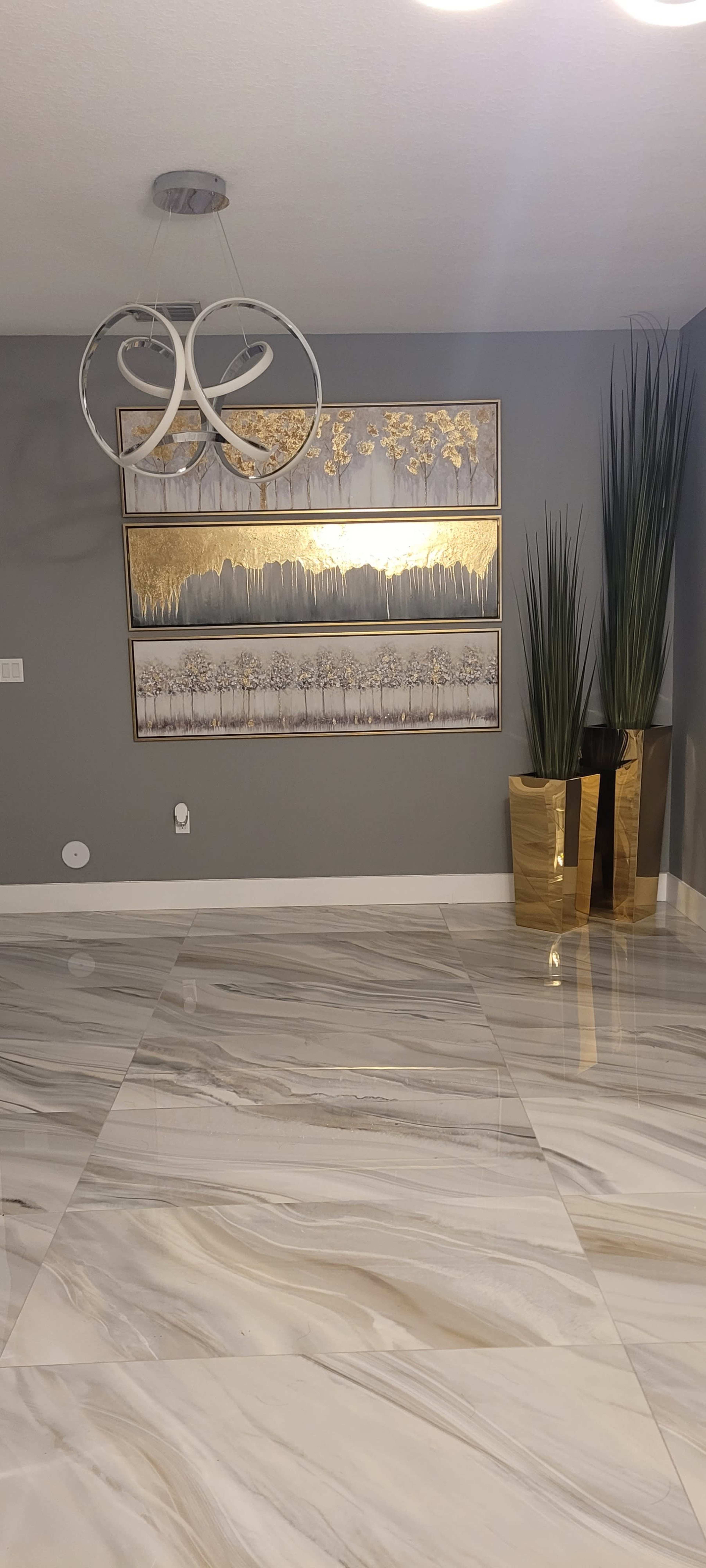 A modern interior space with marble flooring, a gray wall featuring decorative artwork, and two tall plants in gold-colored planters.