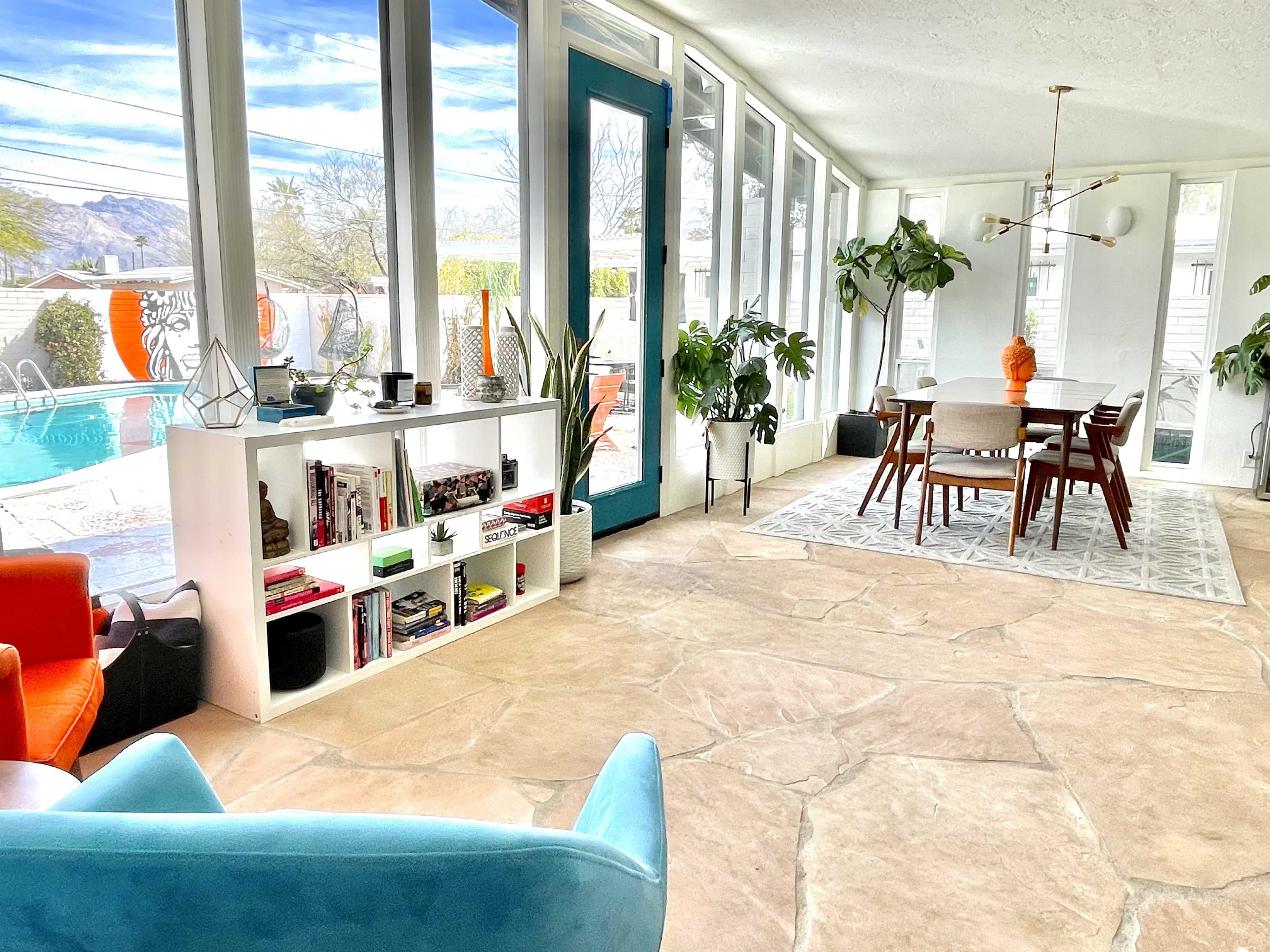 A bright, modern living space with a dining area, a bookshelf, large windows, and a view of an outdoor pool.