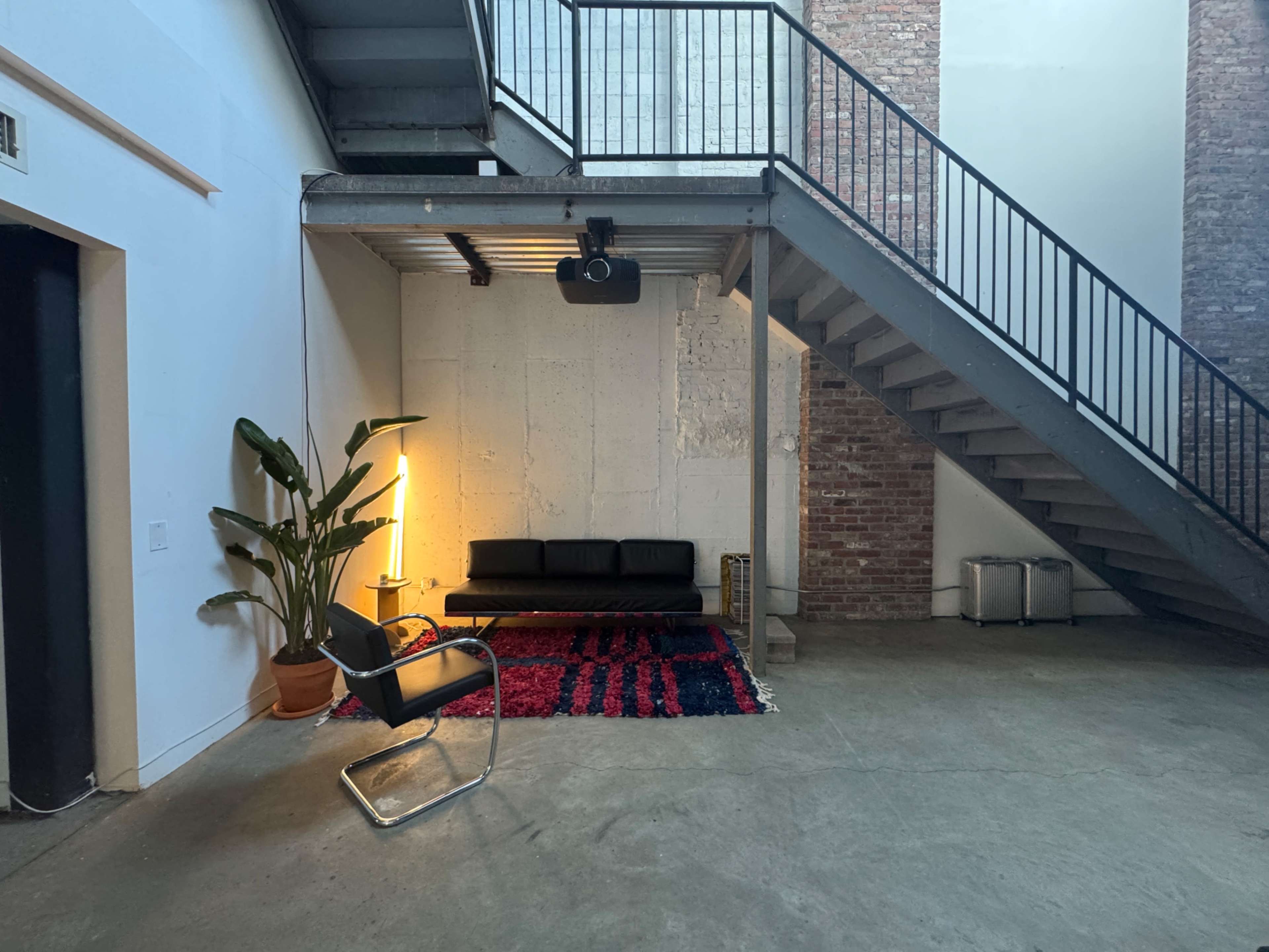 The image shows a spacious interior with a staircase, a black couch, a chair, a potted plant, and a rug on the concrete floor.