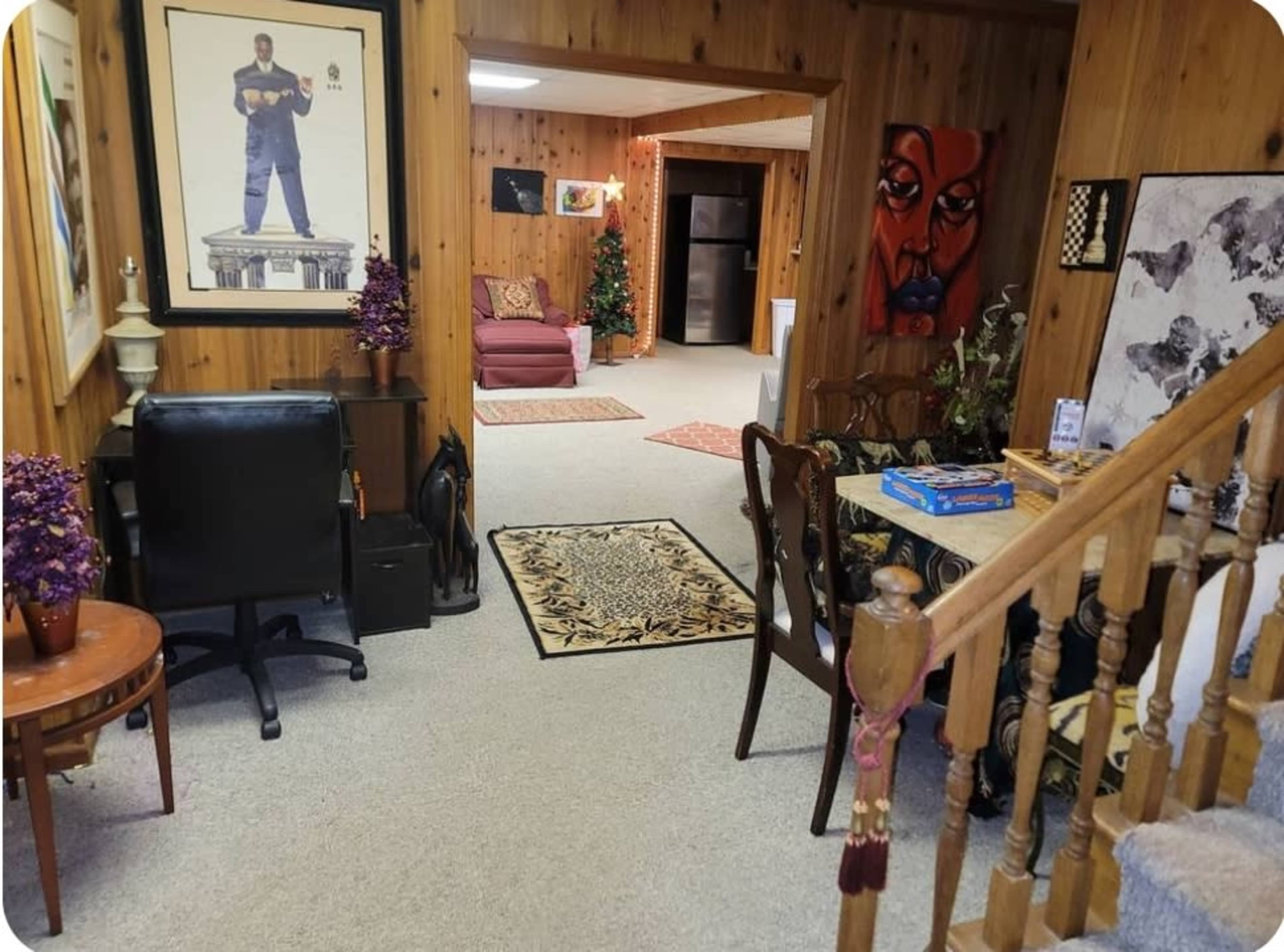 The image shows a cozy basement area featuring wooden paneling, a seating area with a couch, a small table with a game, and various decorations, including artwork and a Christmas tree.