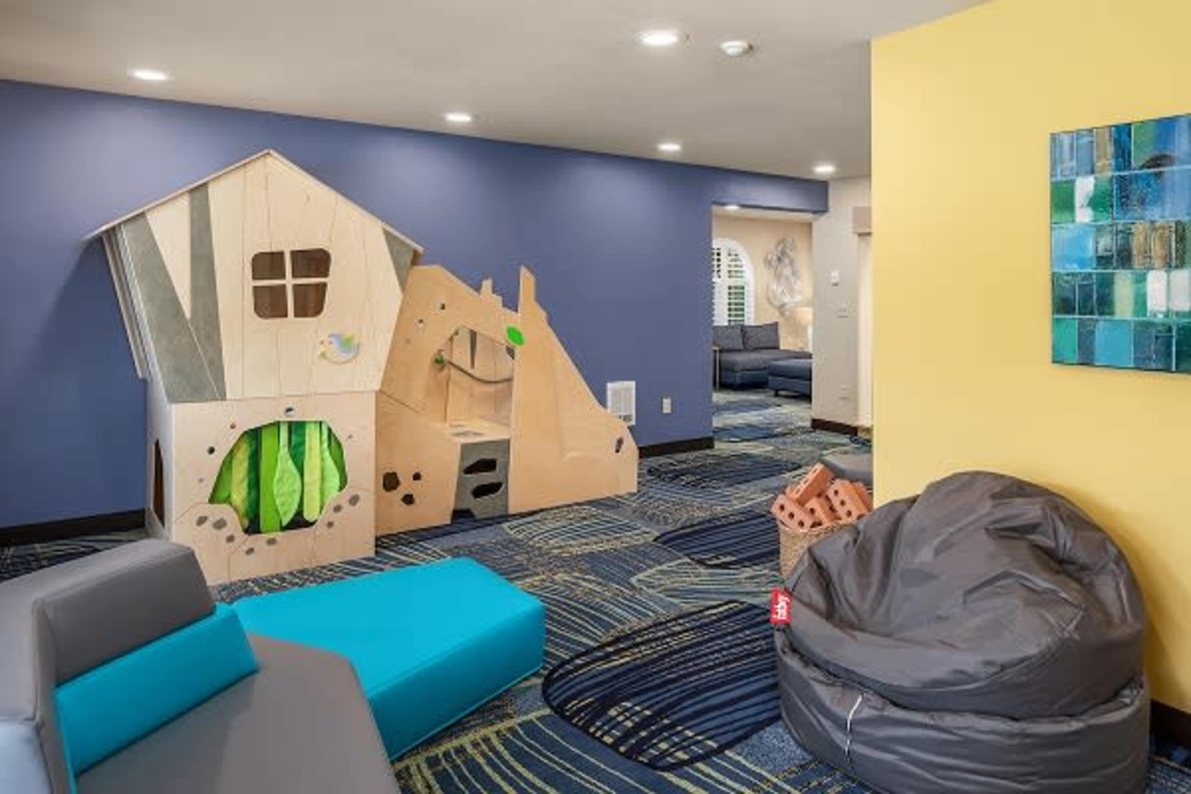 A children's play area featuring a wooden playhouse structure, bean bag chairs, and blue carpet with a patterned design.
