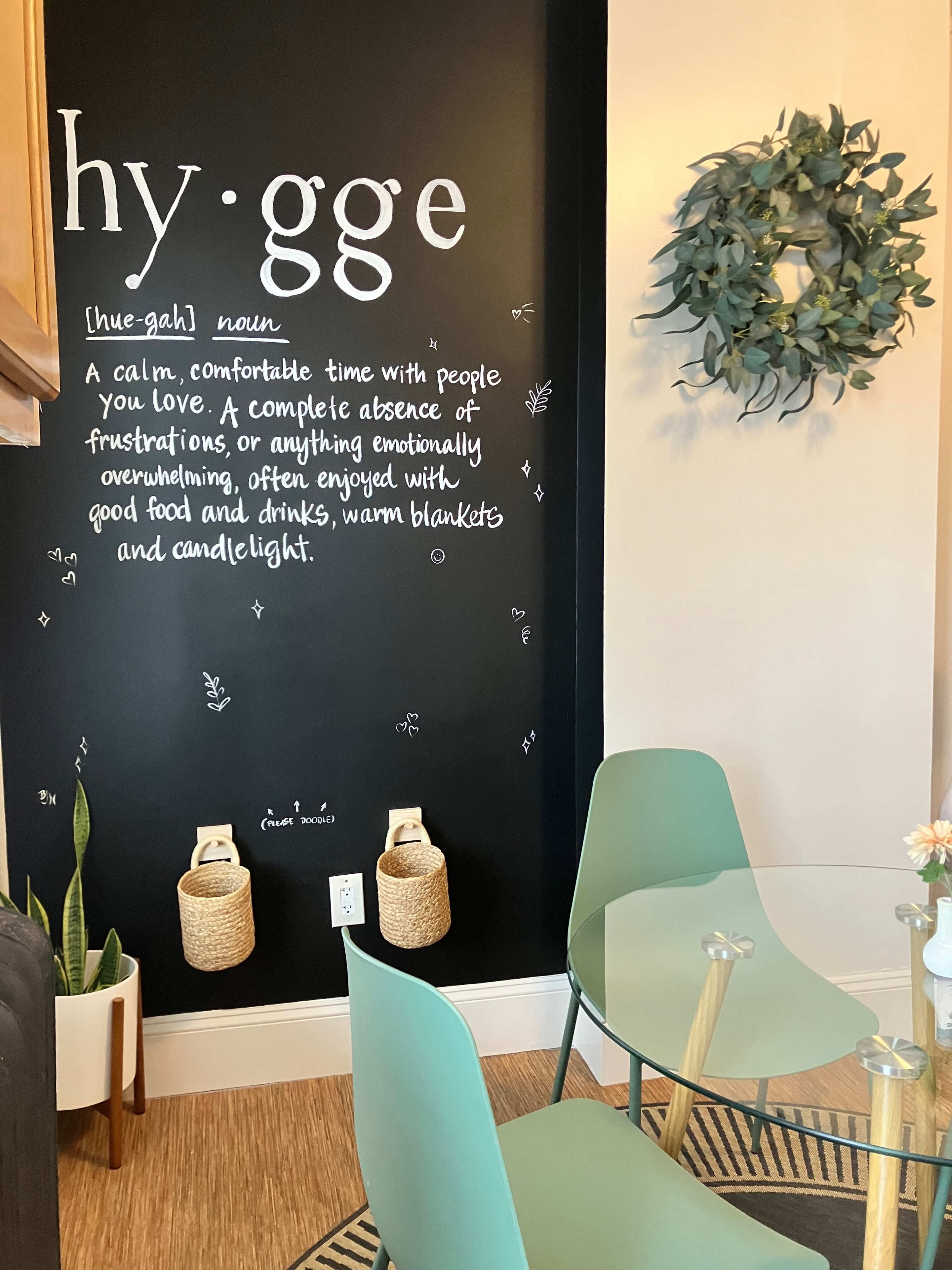 The image shows a cozy corner of a room featuring a black wall with a definition of "hygge" written in white, accompanied by a wreath and a circular glass table with green chairs.