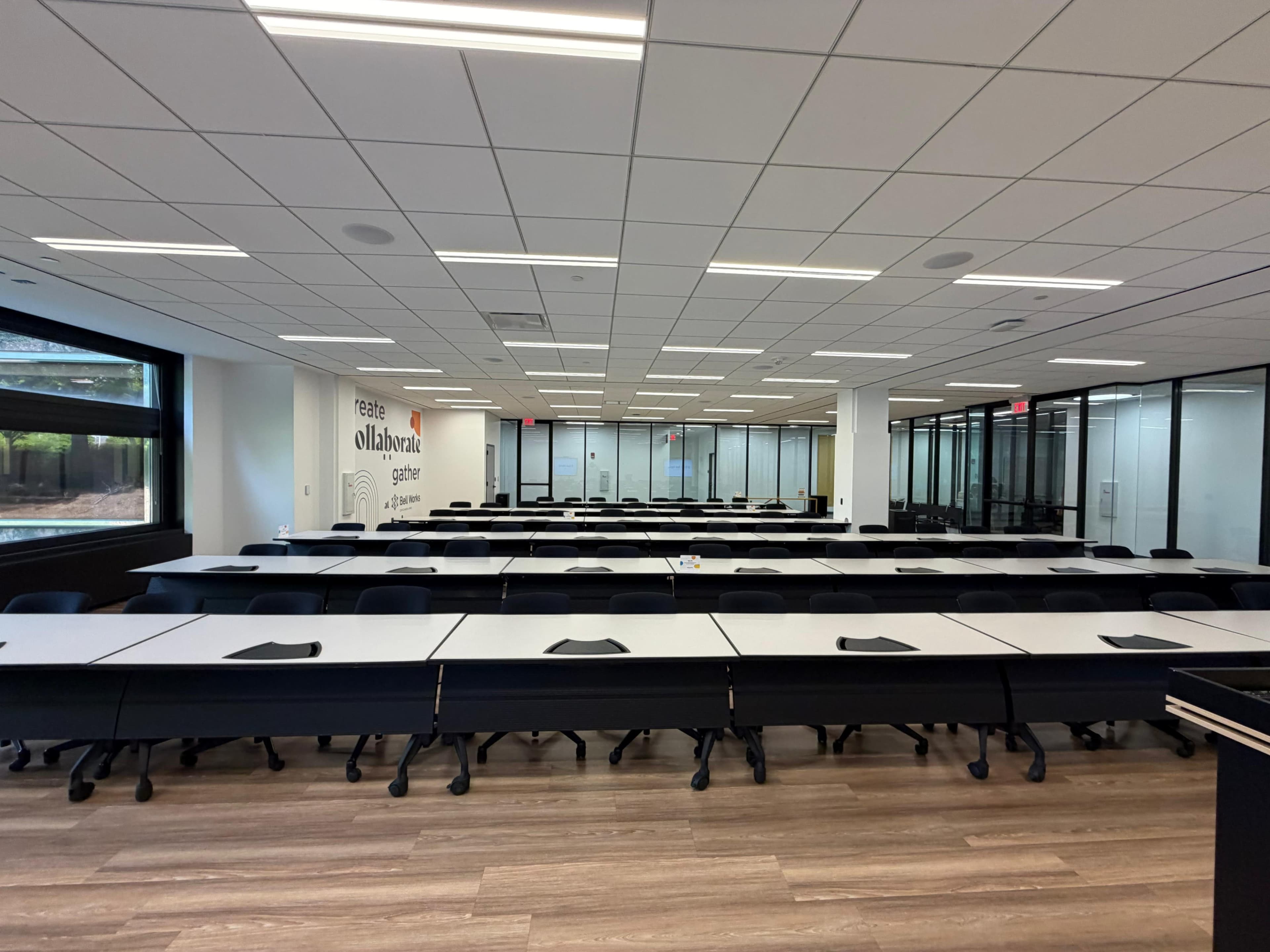 The image shows a modern classroom or conference room with rows of desks and chairs, large windows, and glass walls, creating an open and spacious environment.
