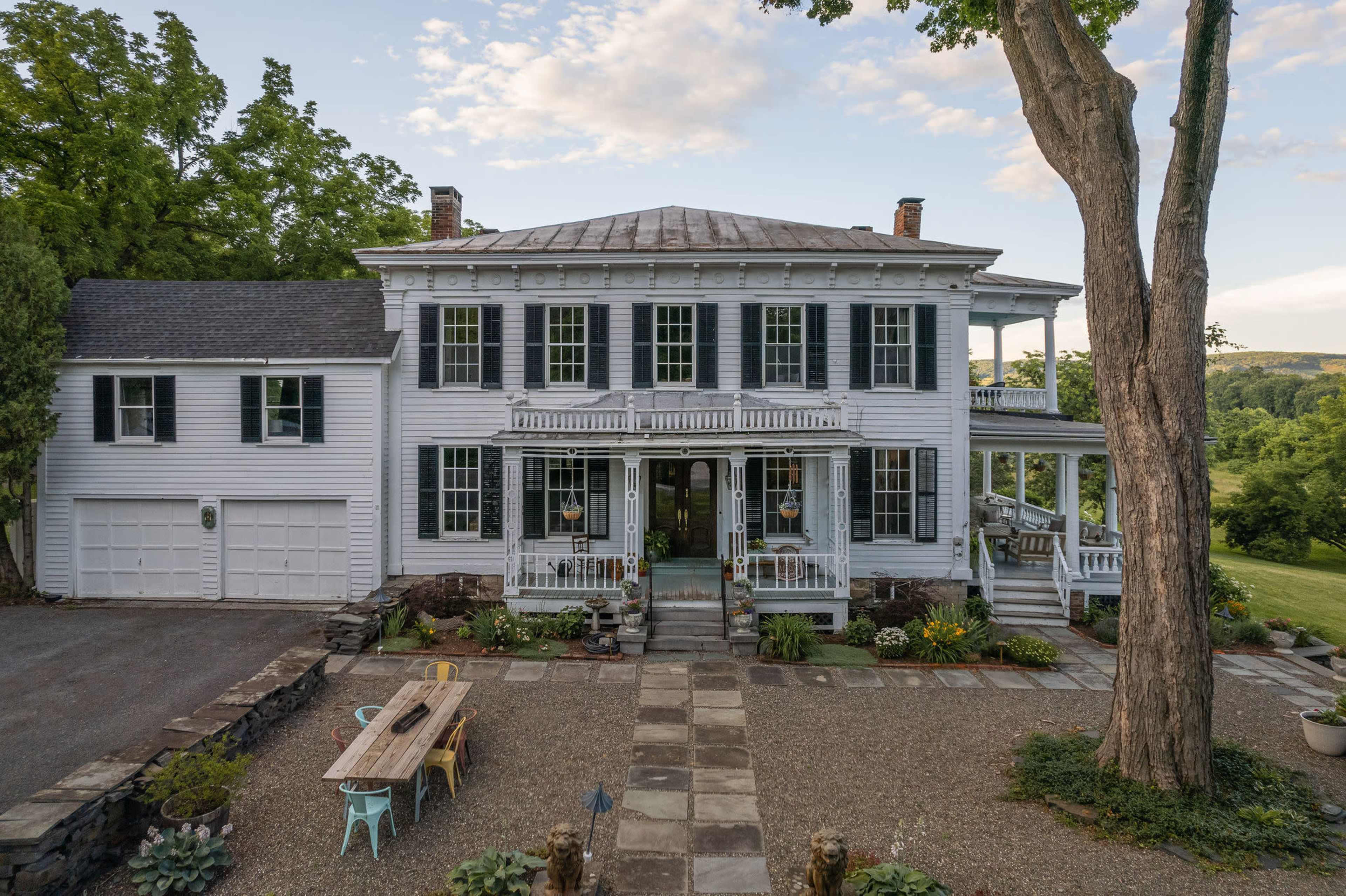 A large, two-story white colonial house with a front porch, surrounded by a gravel courtyard and greenery.