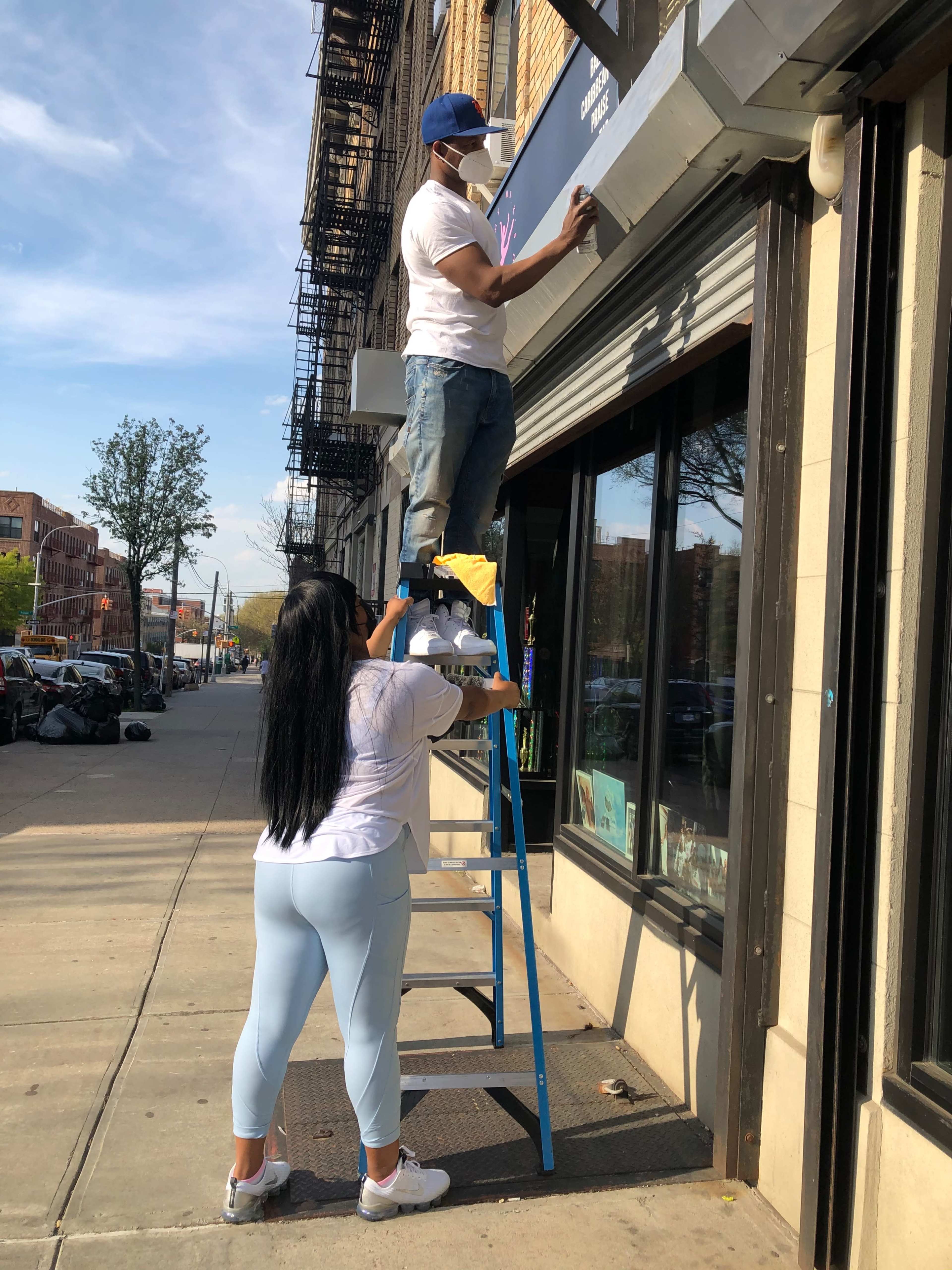 A man stands on a ladder installing a sign above a storefront while a woman assists by holding the ladder steady.