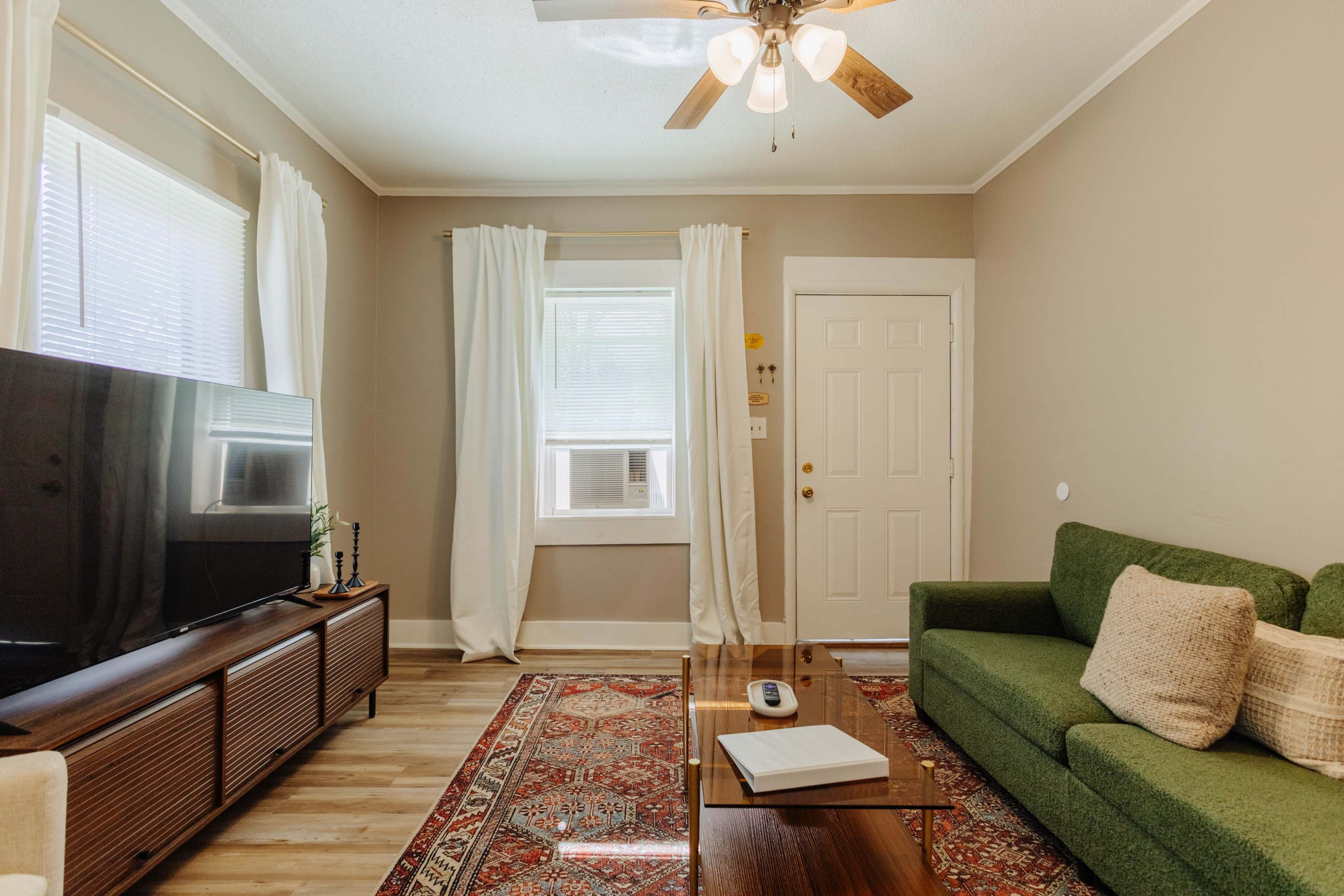 The image shows a cozy living room featuring a green sofa, a wooden coffee table, a television unit, and light-colored curtains, with a door visible in the background.