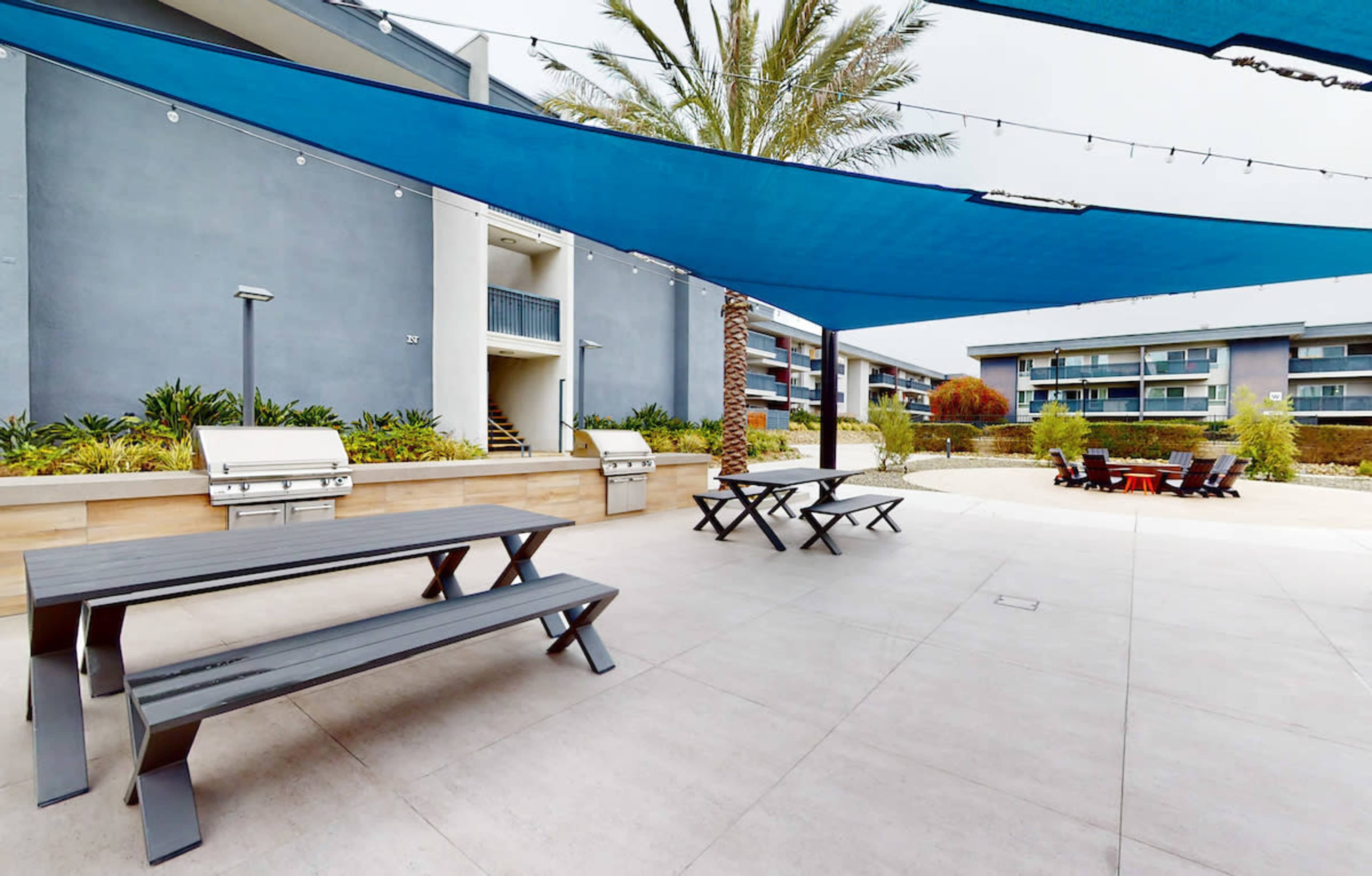 A landscaped outdoor area with shaded picnic tables, outdoor grills, and seating near a building complex.
