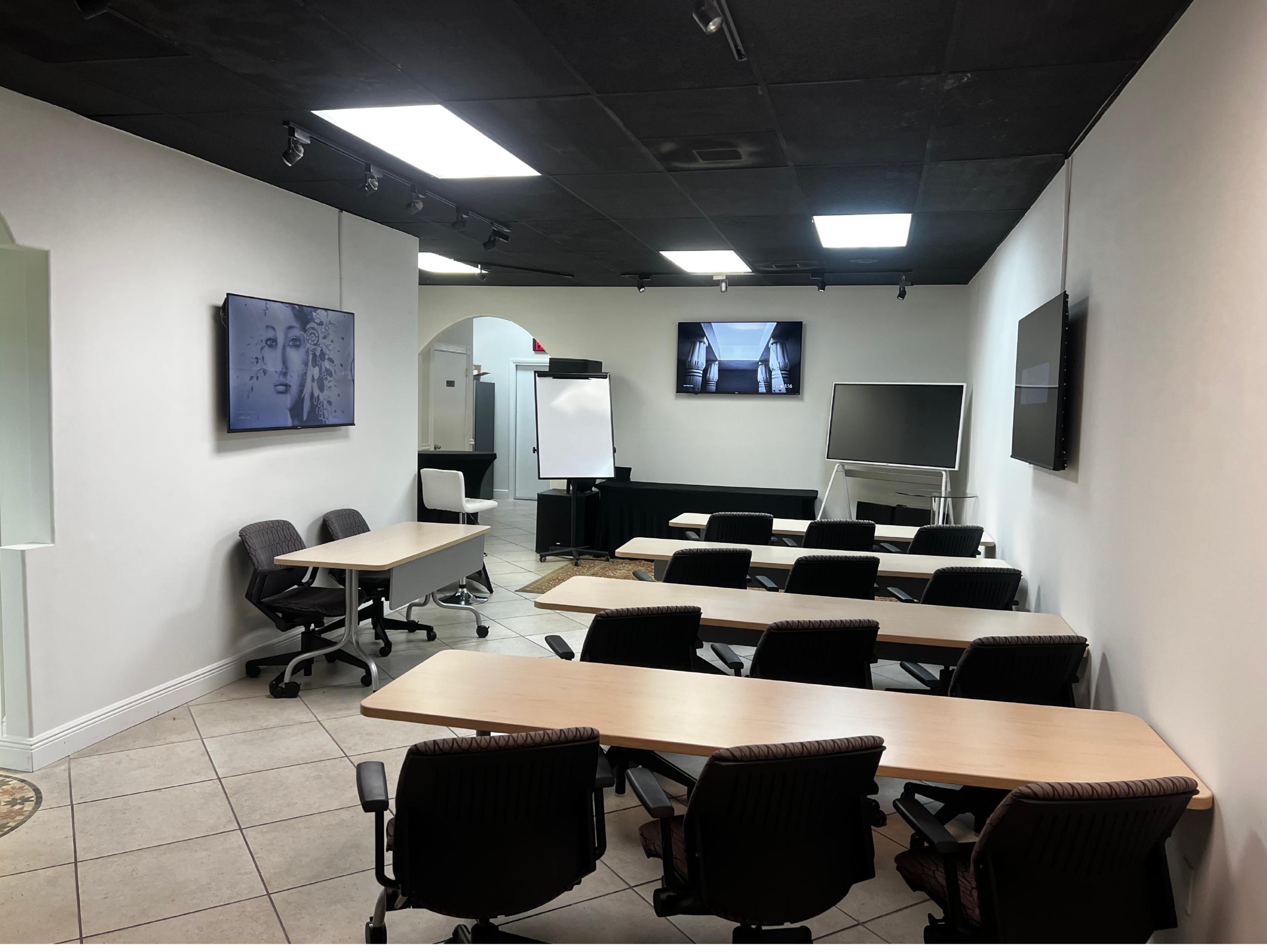 The image shows a small conference room equipped with several tables, chairs, and displays, with a whiteboard at the front.