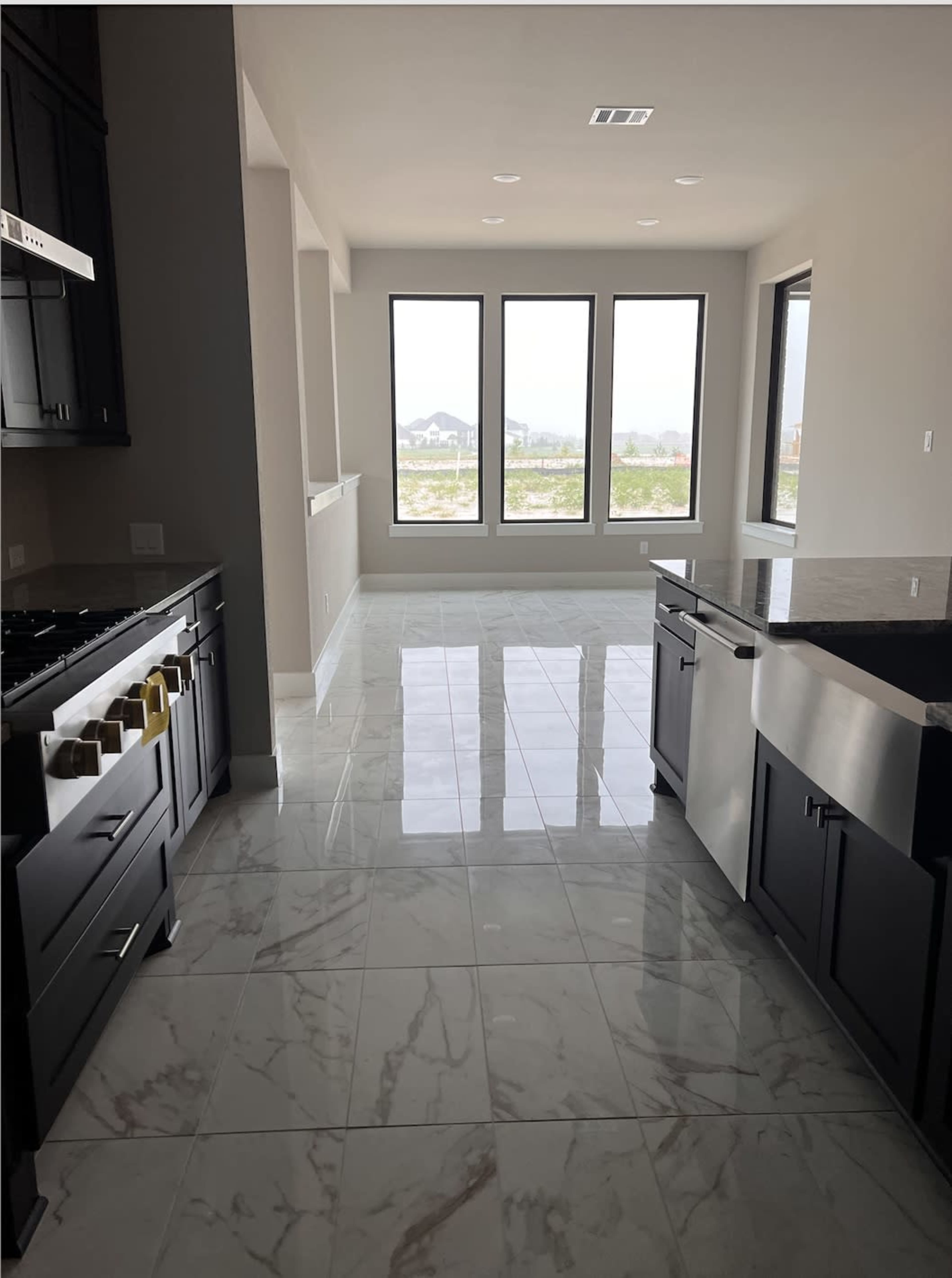 The image shows a modern kitchen with dark cabinetry, a large island, and glossy marble floor tiles leading to a wall of windows that overlook an outdoor view.