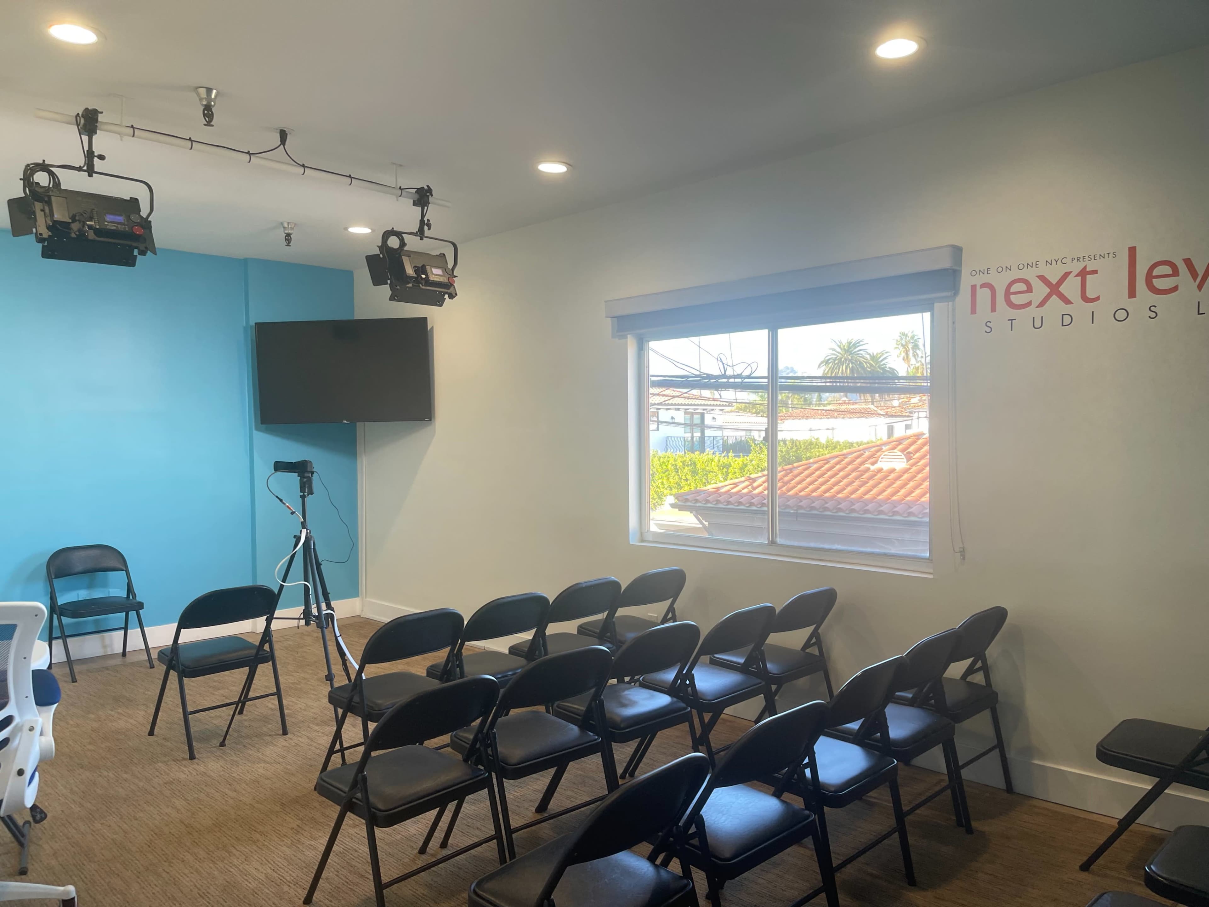 The image shows a small room arranged for a presentation, featuring folding black chairs, a mounted TV, and two overhead lights, with a window that overlooks a rooftop.