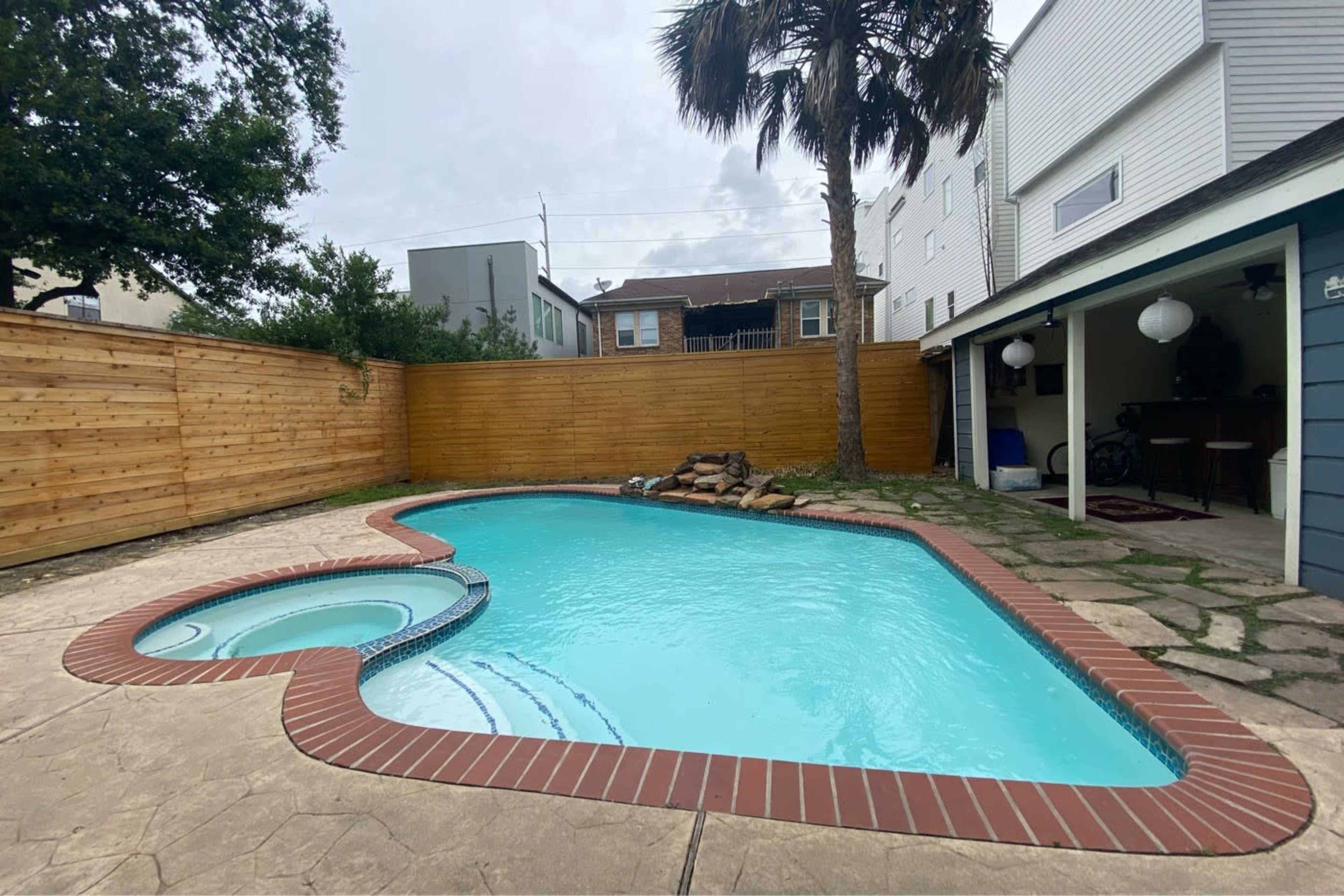 A backyard pool in a heart shape with a spa feature, surrounded by a wooden fence and a palm tree.