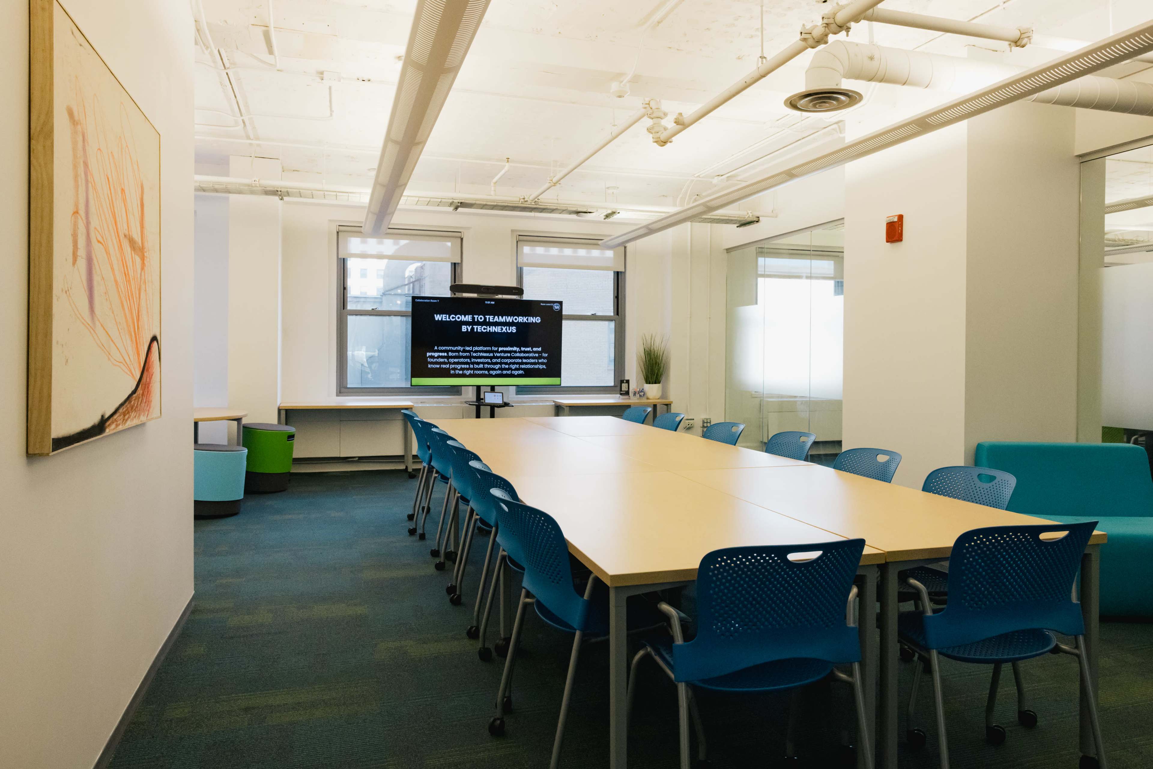 A conference room features a long table surrounded by blue chairs, with a screen displaying a welcome message on one wall.