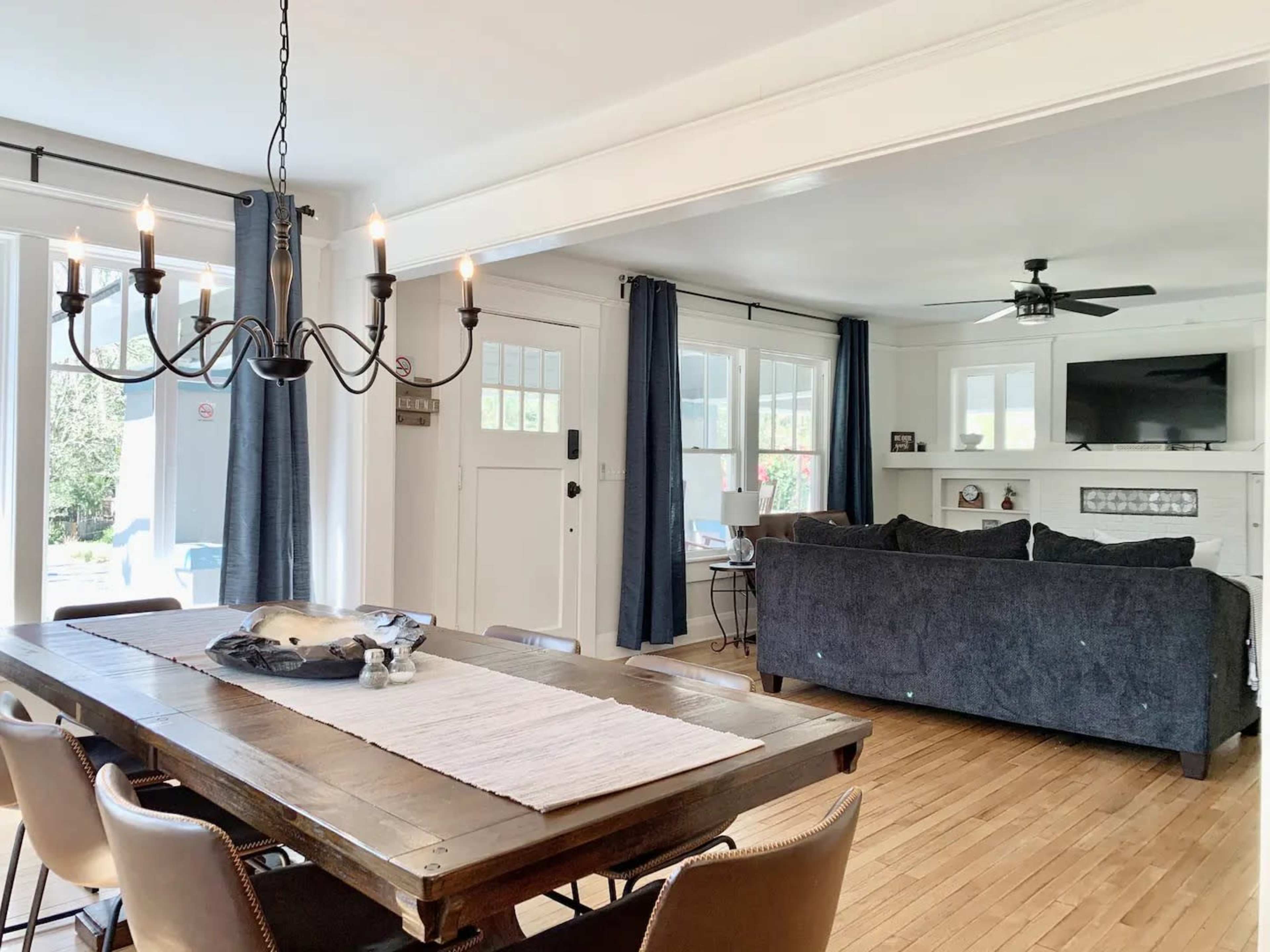 A dining area features a wooden table with chairs, a chandelier overhead, and an open view into a living space with a couch and television.
