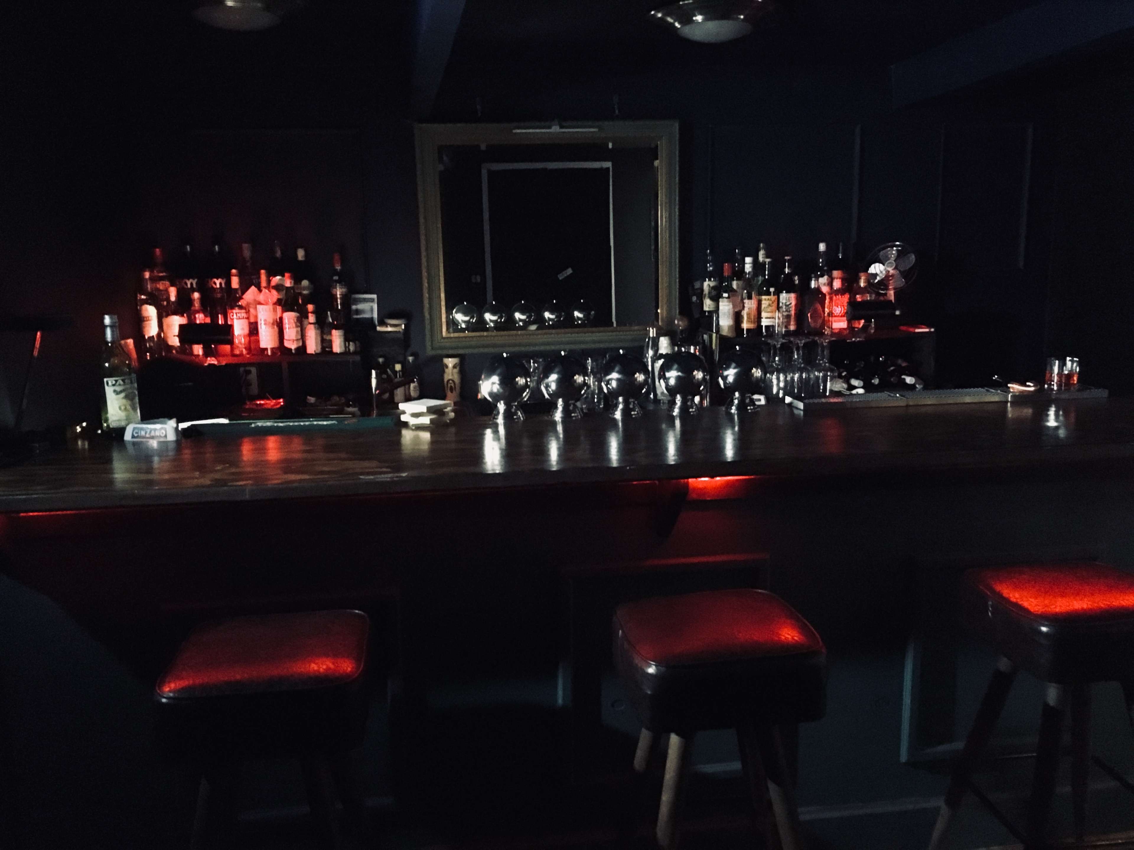A dimly lit bar with a mirrored back, various bottles displayed on shelves, and three stools against the counter.