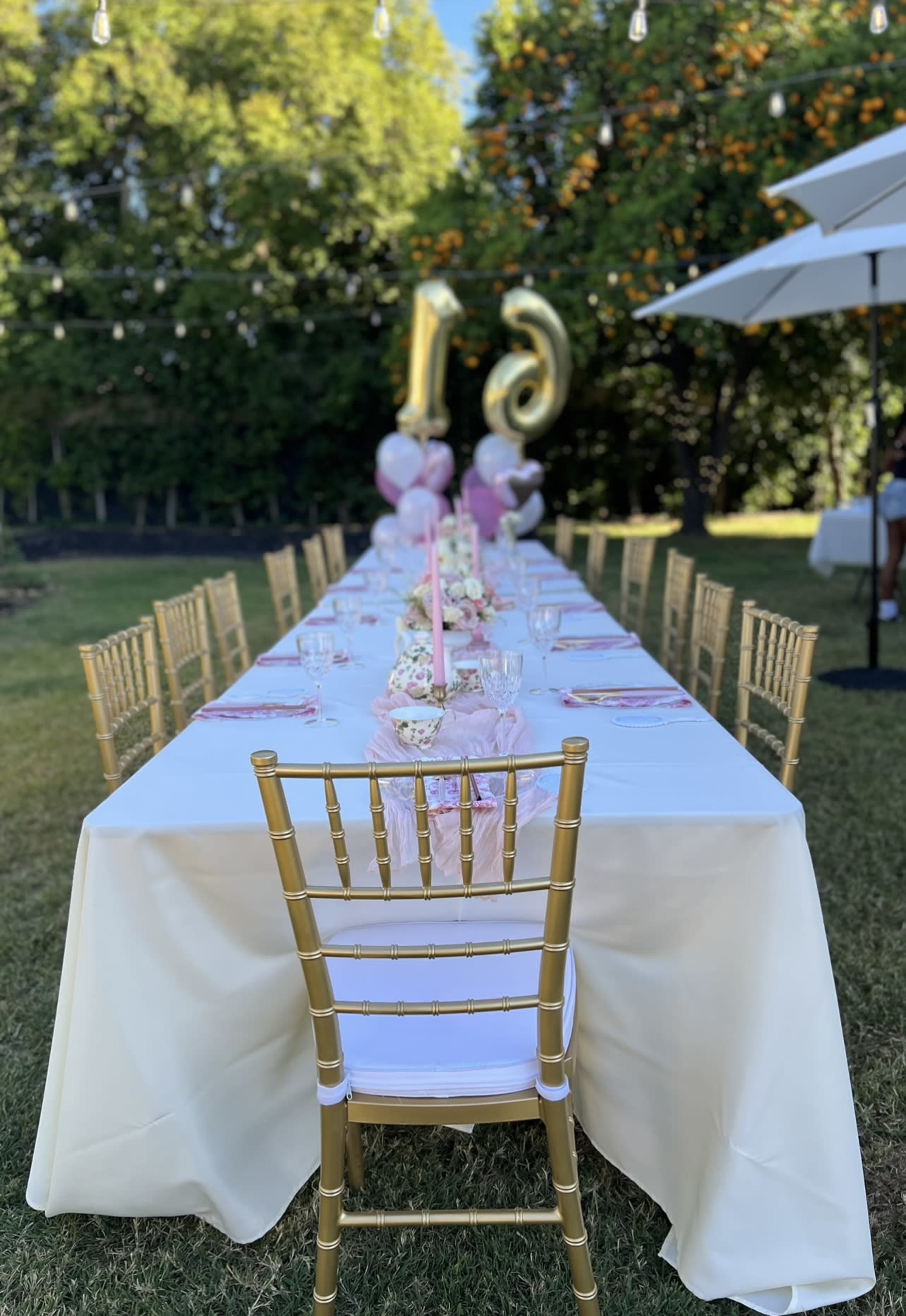 A long, elegantly set outdoor dining table is decorated with flowers and balloons, featuring gold chairs under a clear blue sky.