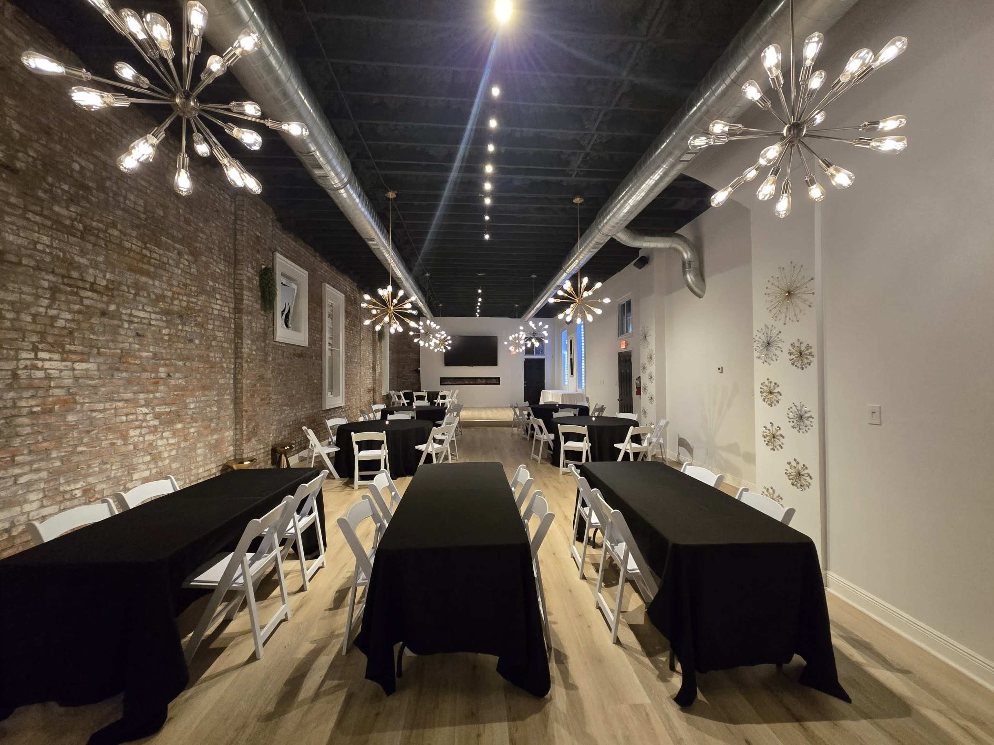 The image shows a spacious event room with long tables covered in black tablecloths, surrounded by white folding chairs and illuminated by modern chandelier lights.