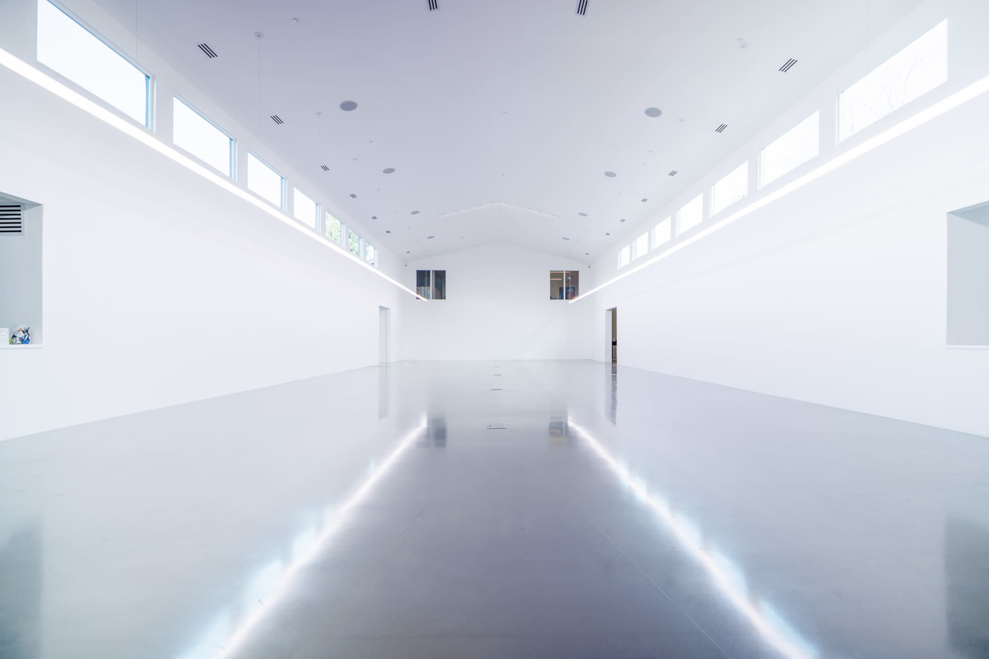 The image shows a large, empty gallery space with white walls and a polished floor reflecting light.