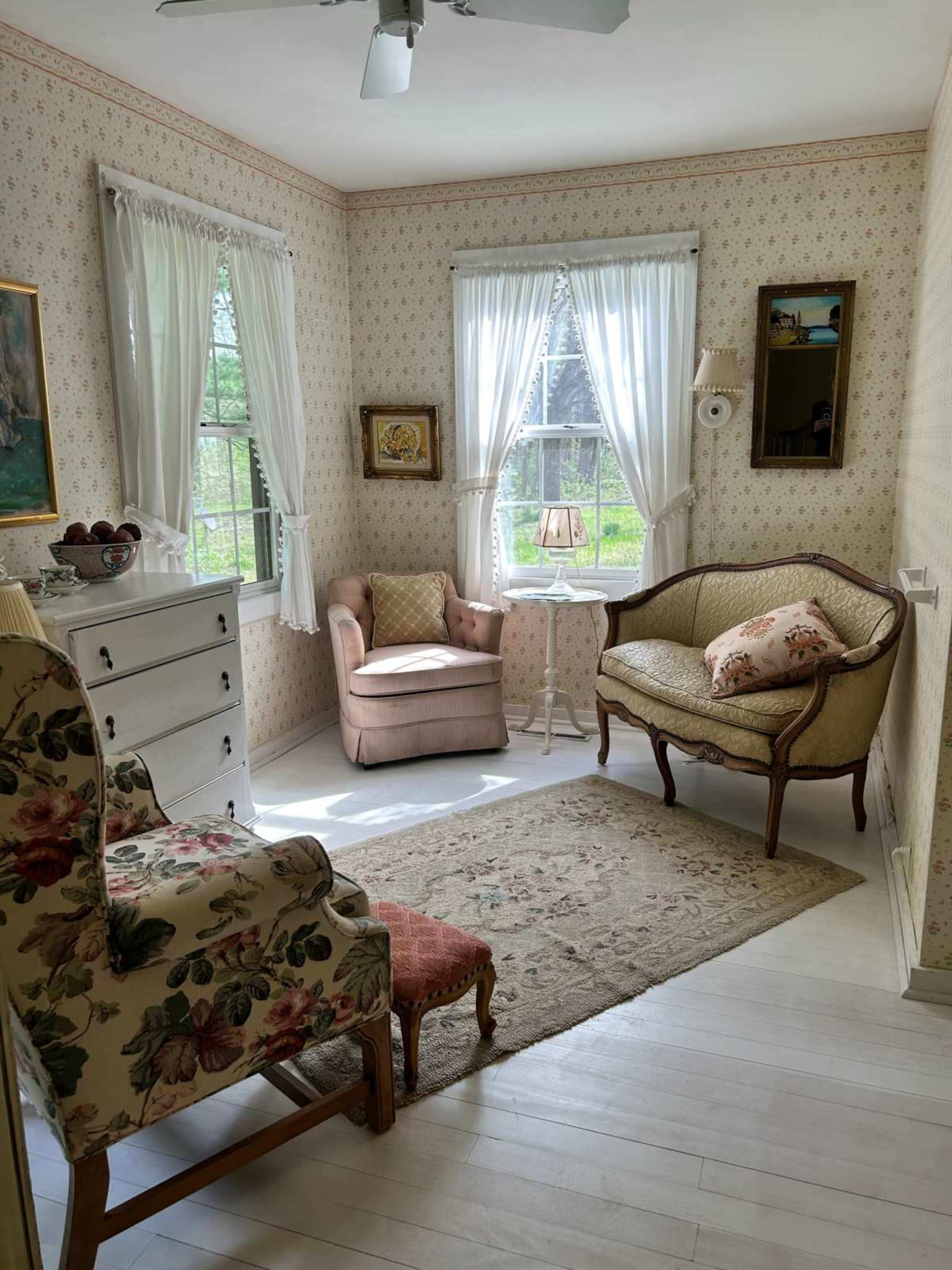 The image depicts a cozy, well-lit room featuring a pink upholstered armchair, a vintage-style settee, and floral-patterned decor, with large windows allowing natural light to enter.