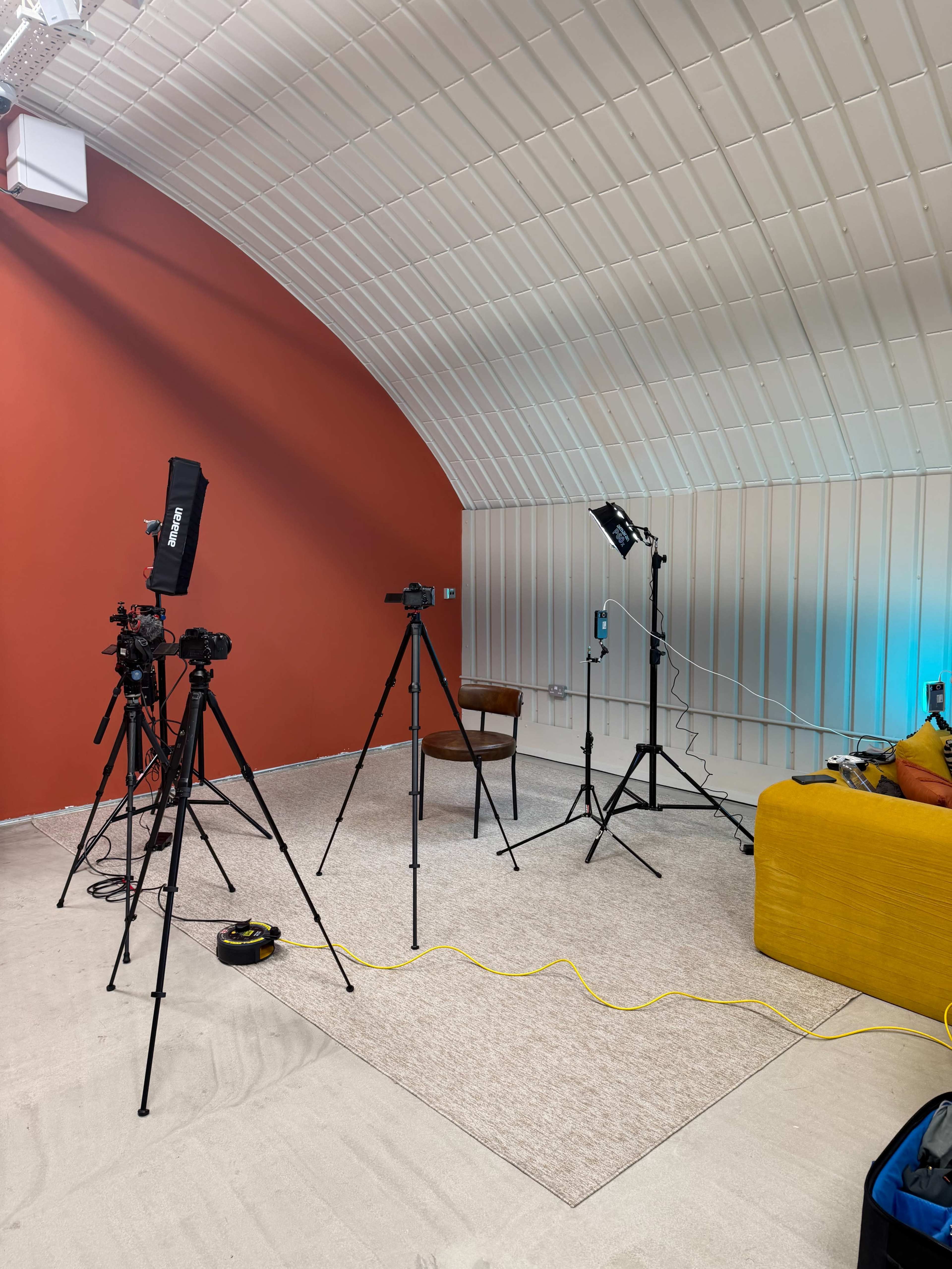 The image shows a studio setup with several tripods holding cameras and lighting equipment, a single chair, and a yellow couch against a red wall.