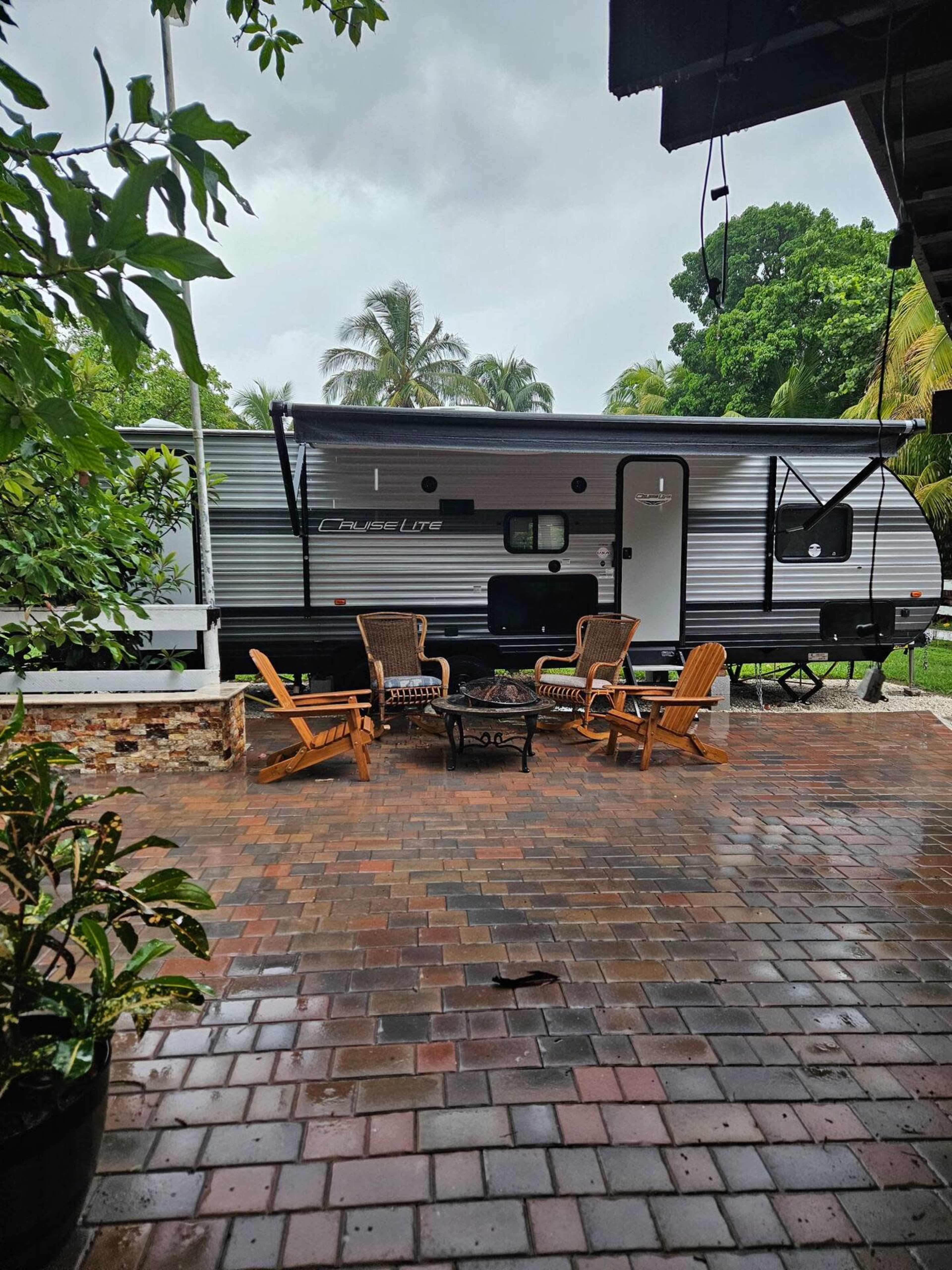 A parked travel trailer sits on a brick patio surrounded by tropical foliage on a rainy day.