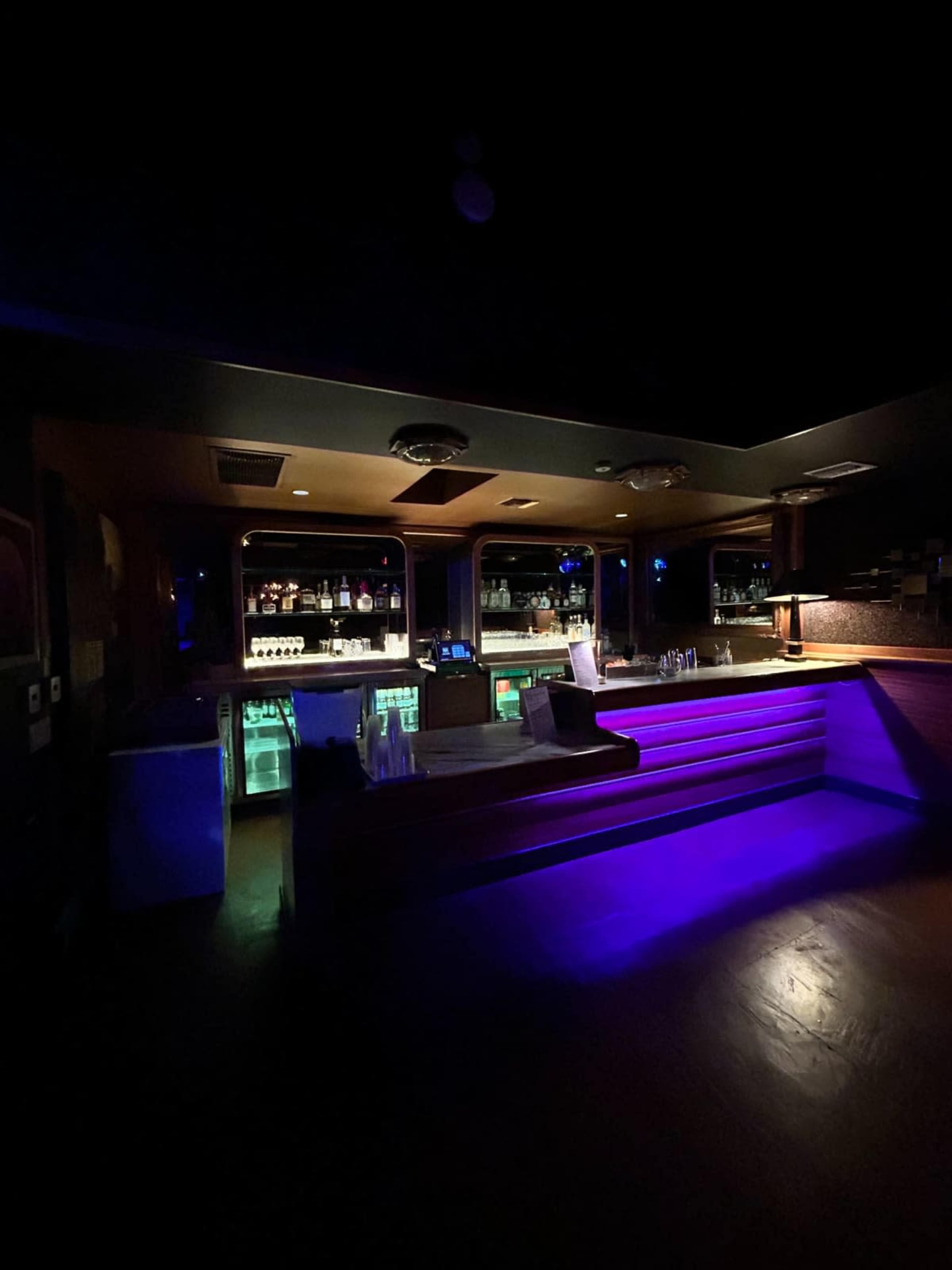 A dimly lit bar features a sleek design with illuminated shelves of liquor and a raised seating area.