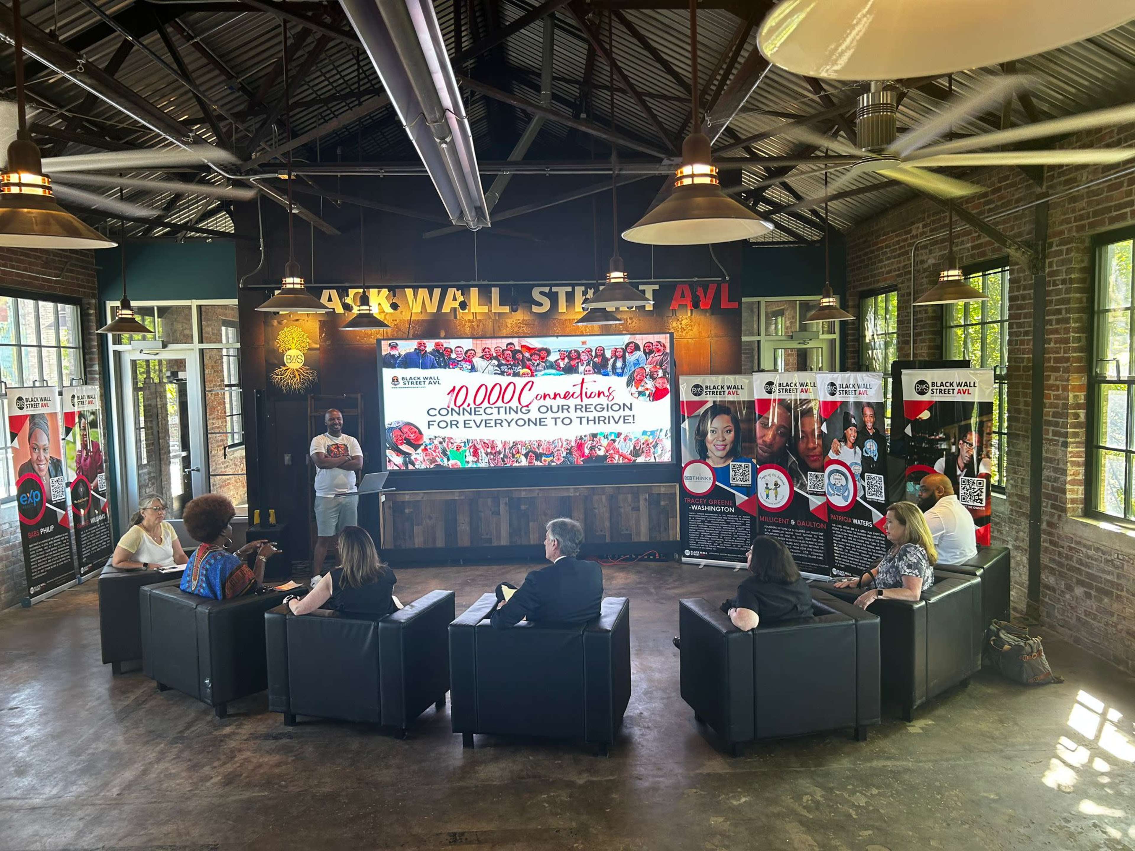 A group of people sits on black couches facing a speaker in a brightly lit room with large banners showcasing community initiatives on the walls.