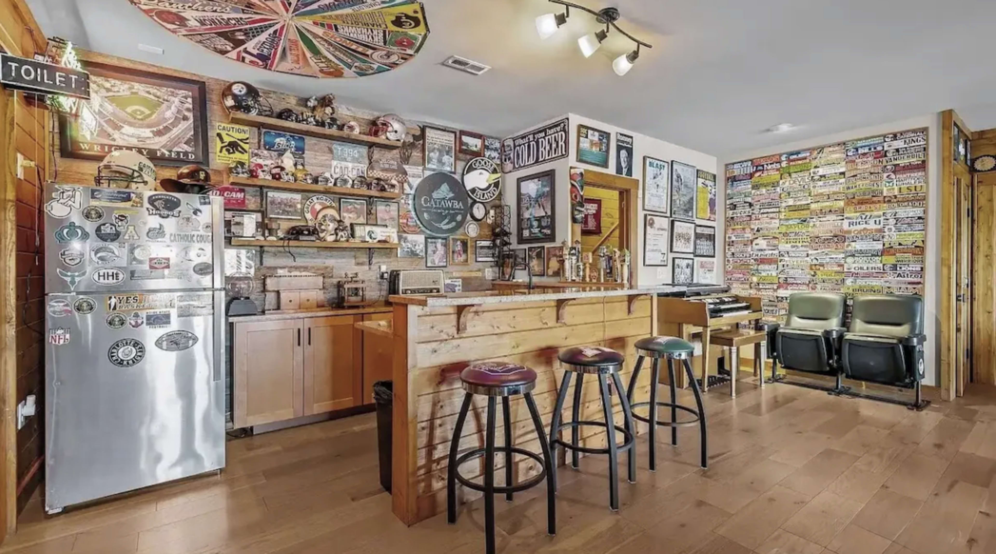 The image shows a rustic bar area with a wooden counter, several bar stools, a decorated wall filled with various signs and posters, and a stainless steel refrigerator.