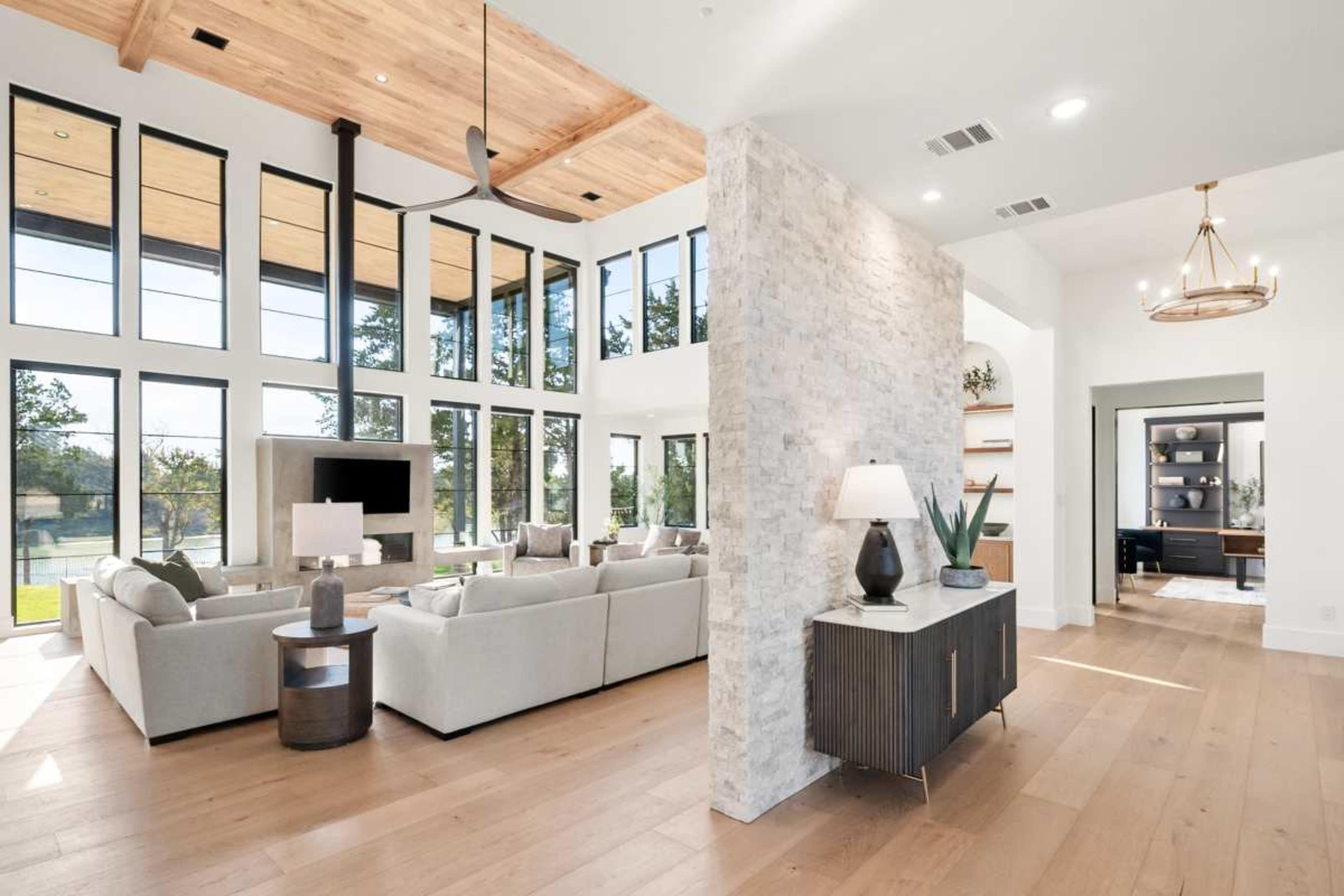 The image features a modern living room with large windows, a stone accent wall, and light hardwood flooring, creating an open and airy space.