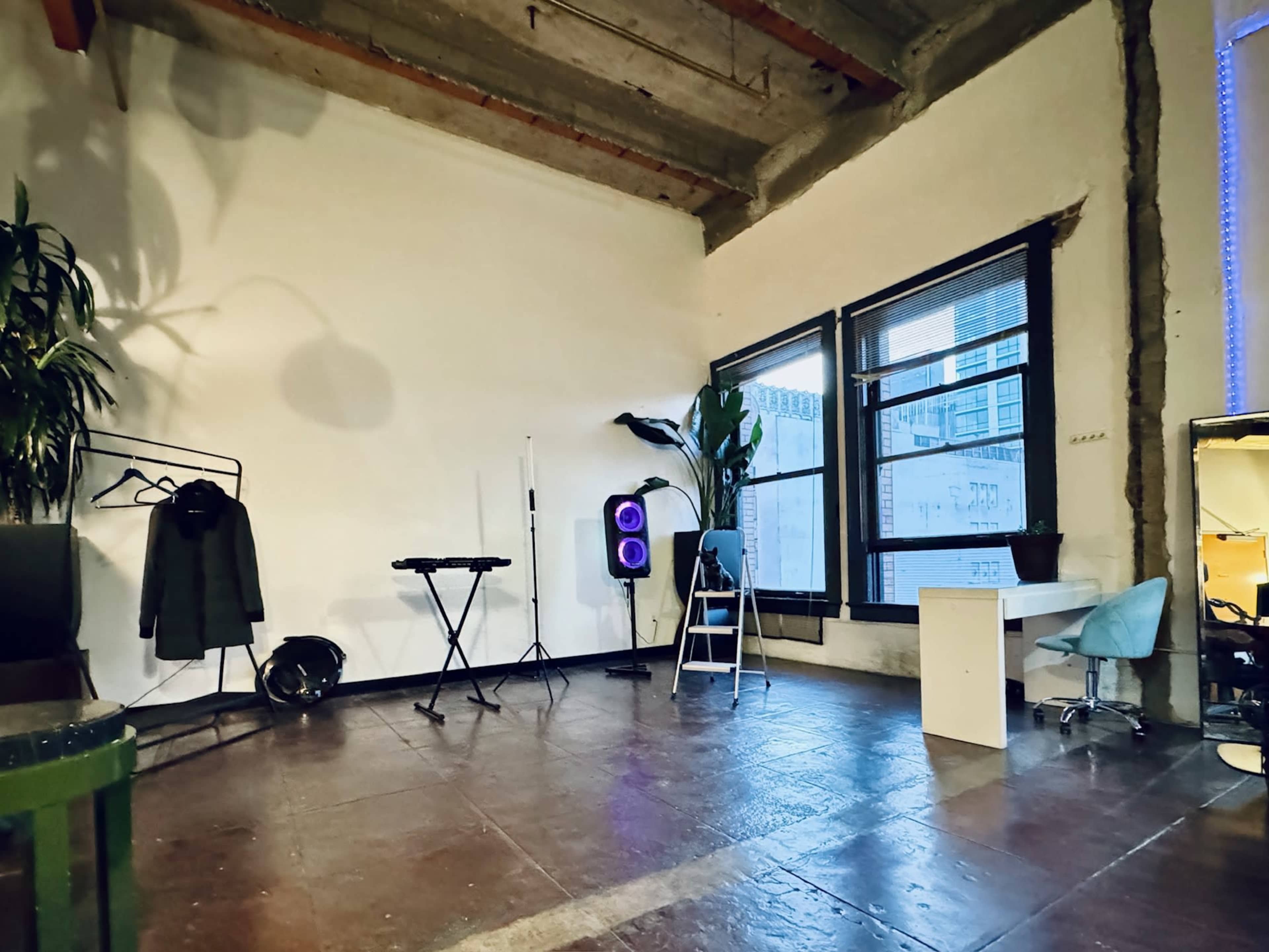A sparse interior space with a keyboard, folding chair, ladder, a potted plant, and a garment rack against a white wall and large windows.
