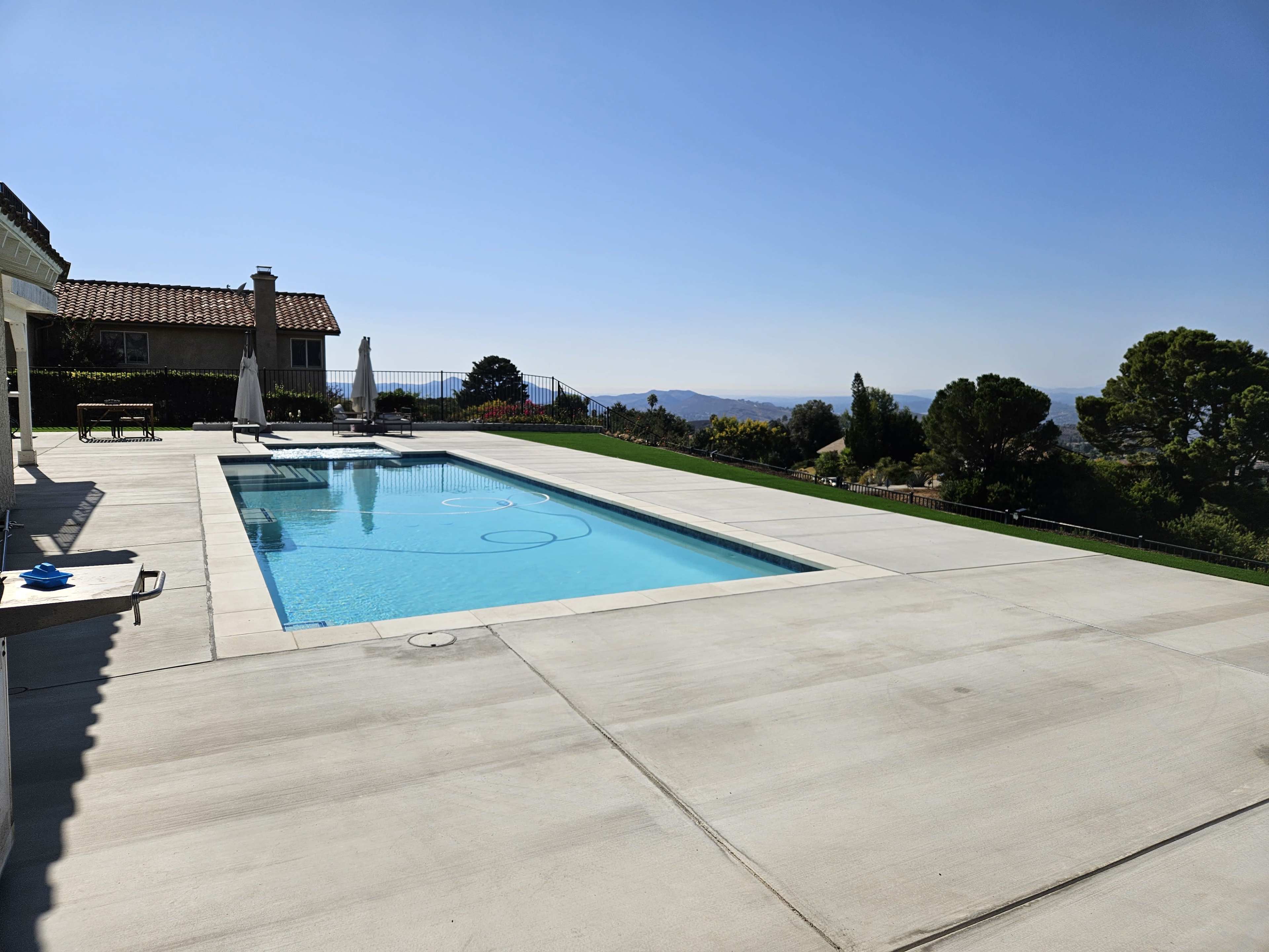 The image features a modern swimming pool surrounded by a concrete patio and a view of distant hills under a clear blue sky.