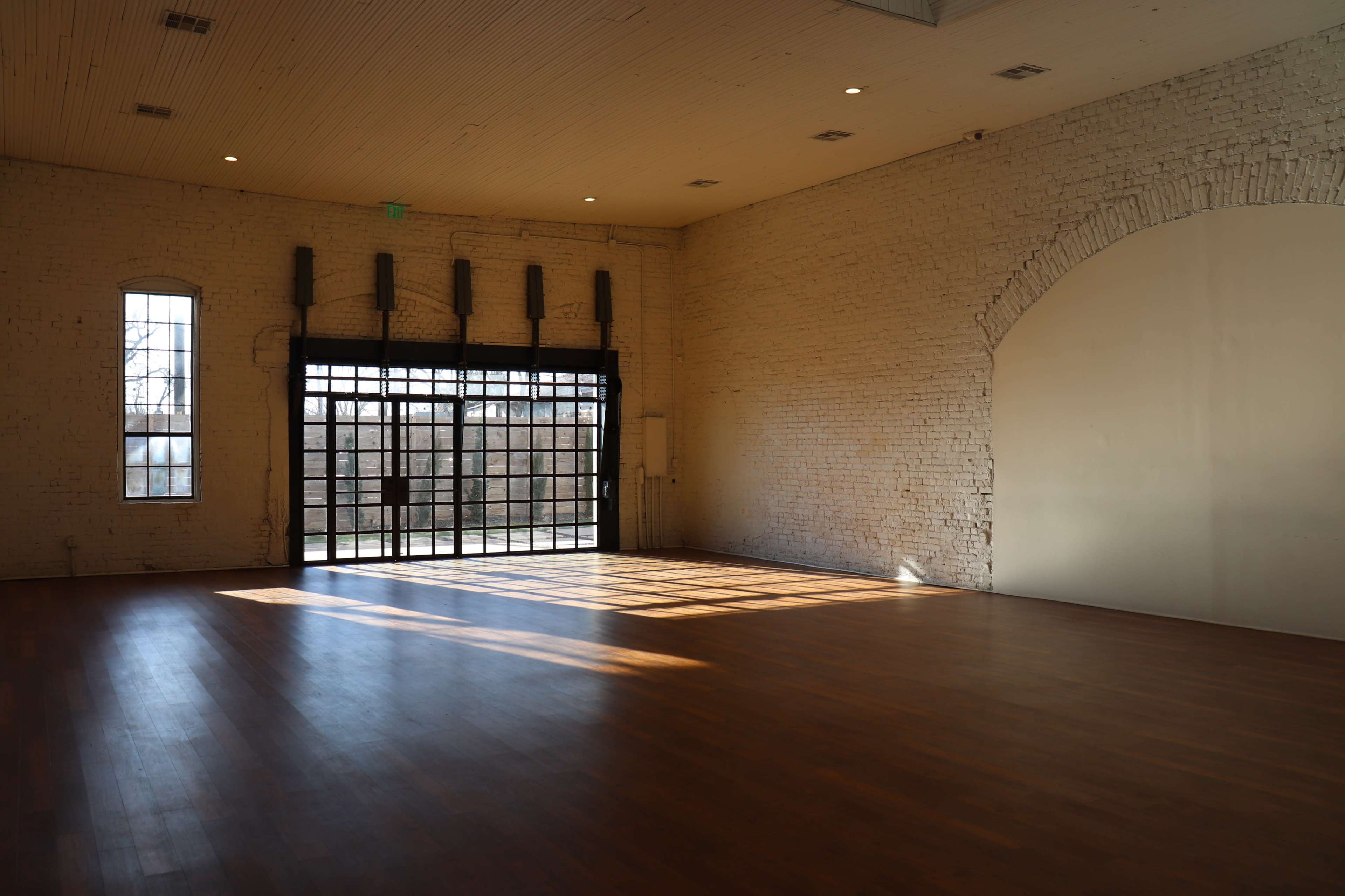 The image shows a spacious, empty room with large windows, wooden flooring, and white brick walls.