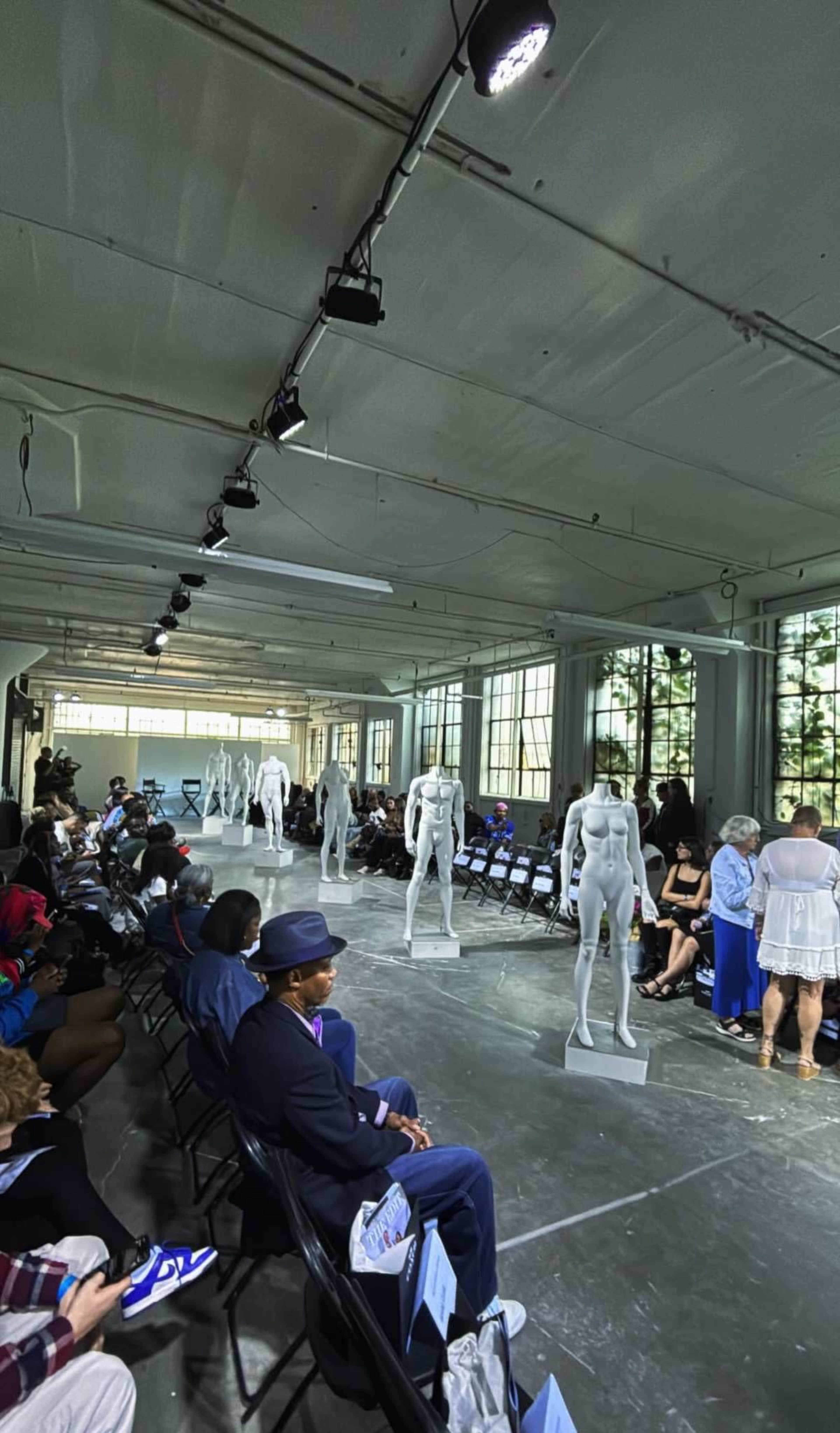 A spacious indoor venue features rows of chairs facing a runway with mannequins positioned at intervals, while an audience sits on either side.