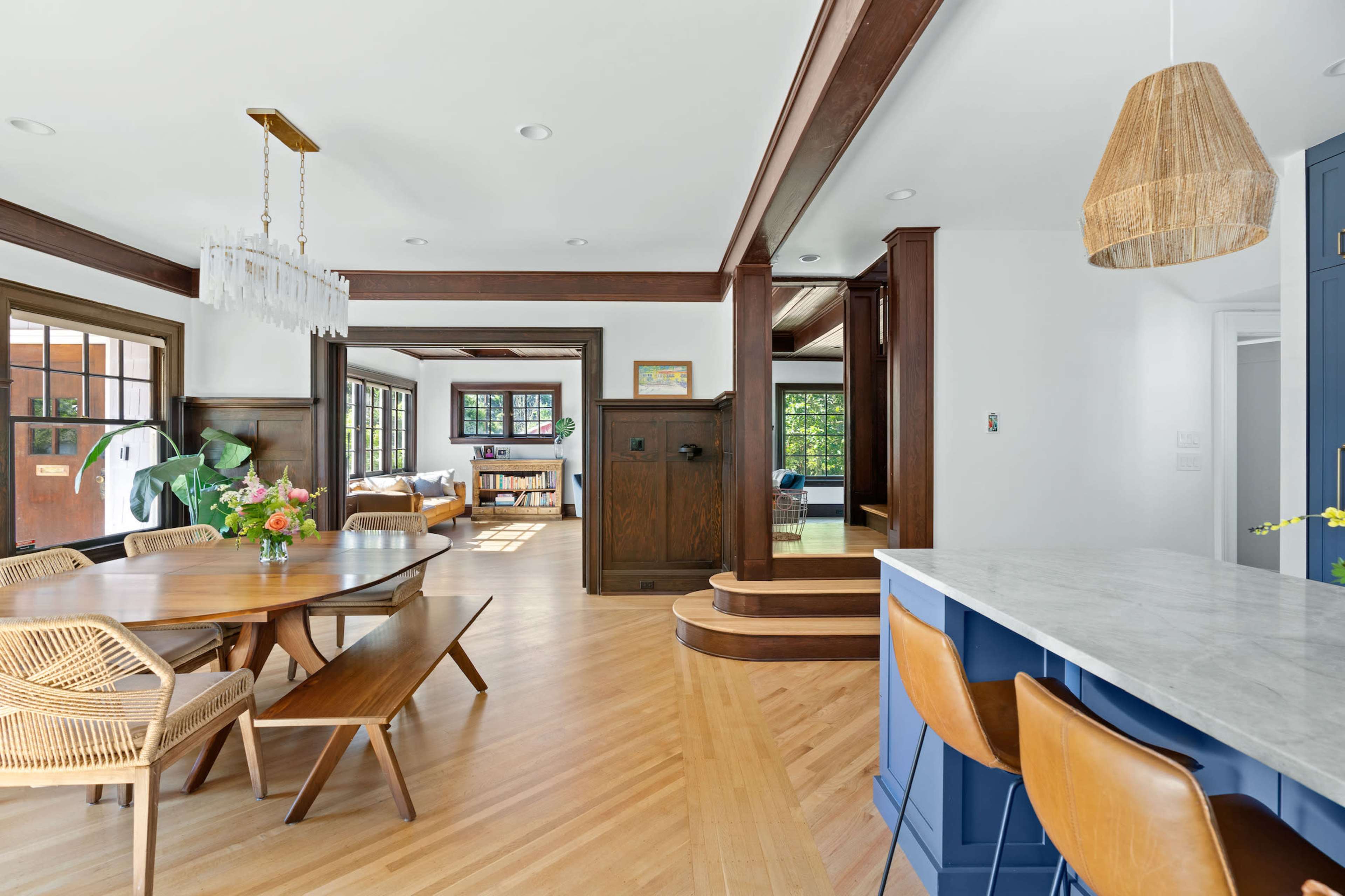 A modern dining area with a wooden table, rattan chairs, and a kitchen with blue cabinetry and a marble countertop, alongside a sunlit living space.