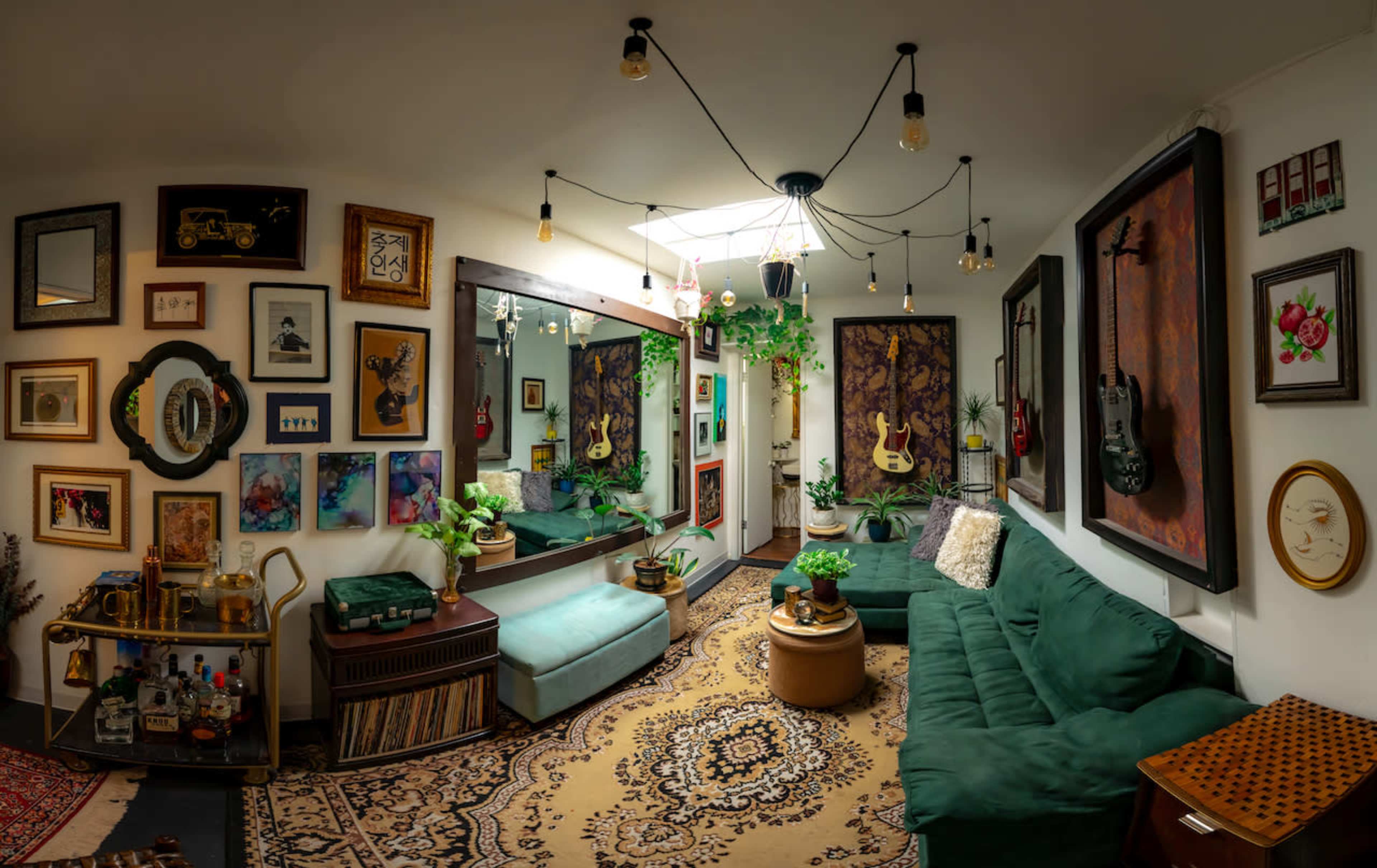 The image shows a stylish living room featuring green sofas, a large mirror, various framed artworks, and musical instruments hung on the walls.