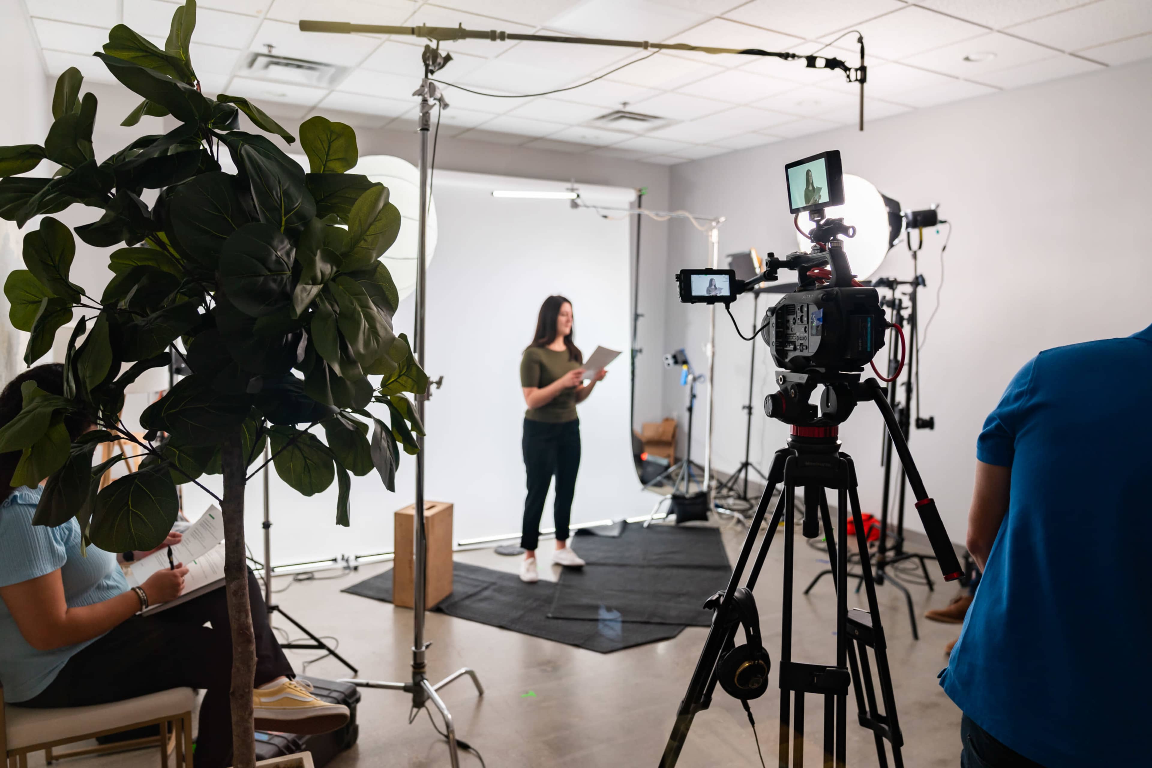 A woman is standing on a black mat in front of a white backdrop while holding a script, surrounded by video equipment and studio lights.
