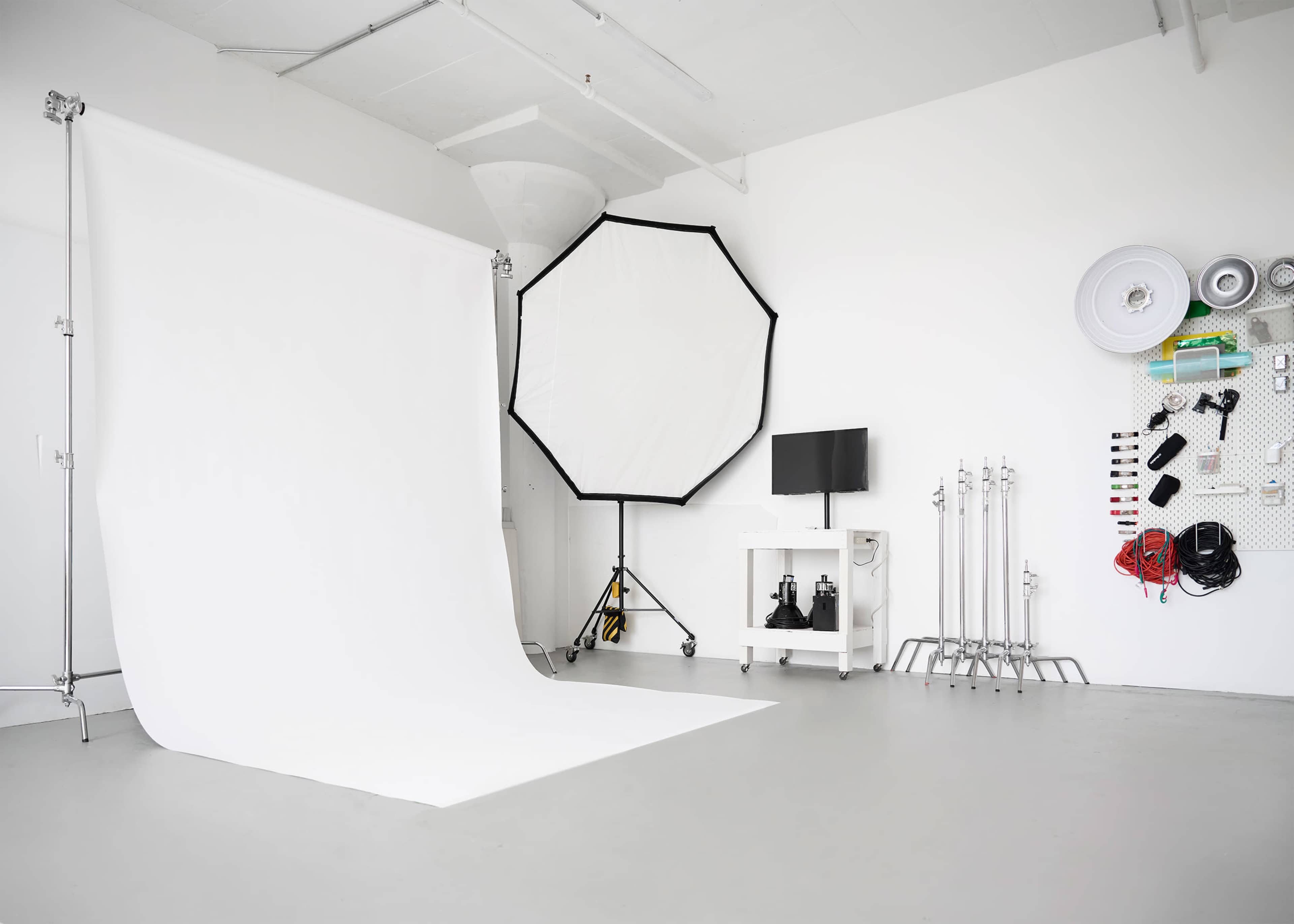 A photography studio featuring a white backdrop, an octagonal softbox, a television on a stand, and equipment organized on a pegboard.