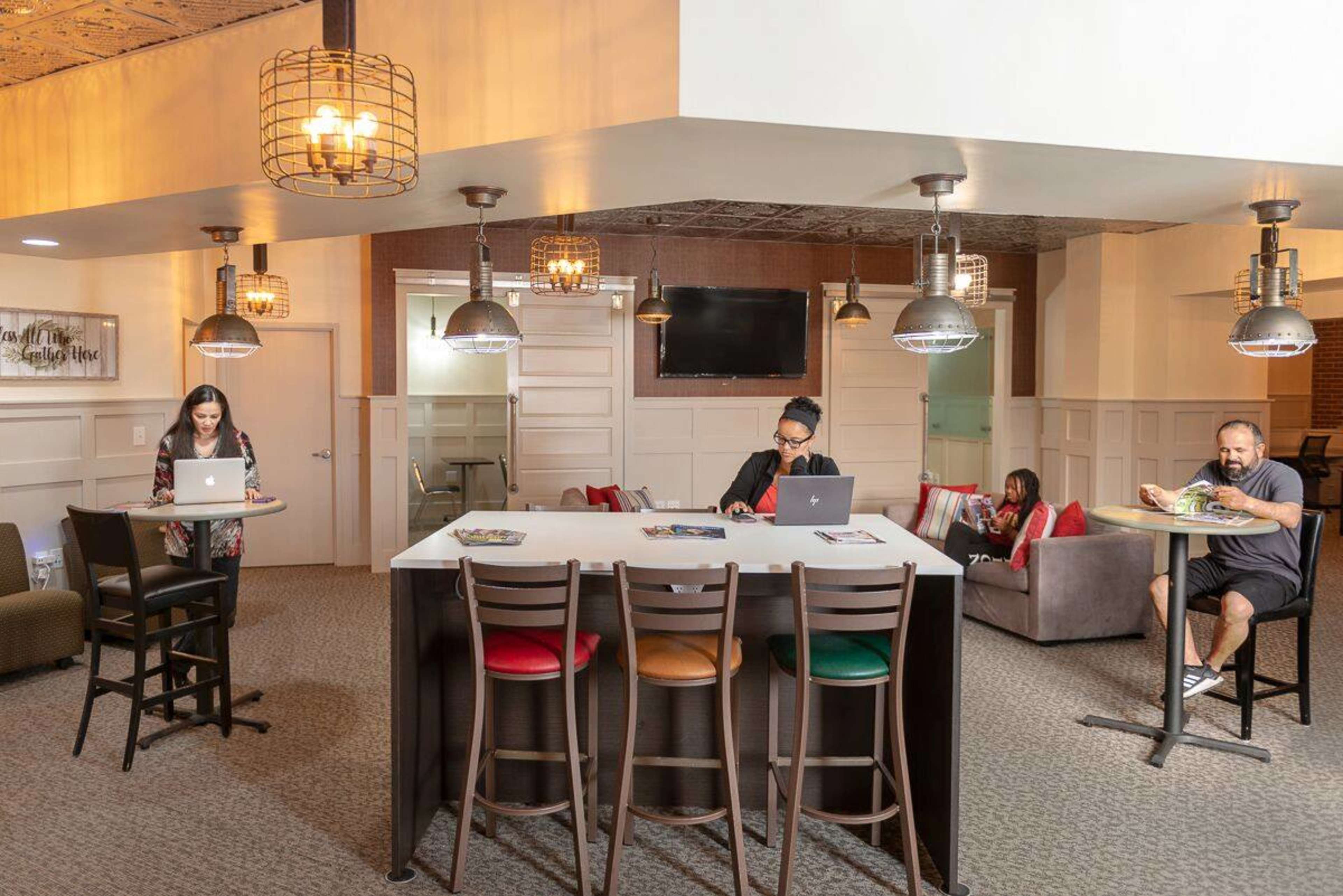 A modern workspace features four individuals using laptops while seated at a bar-height table and nearby seating in a well-lit area with decorative pendant lighting.